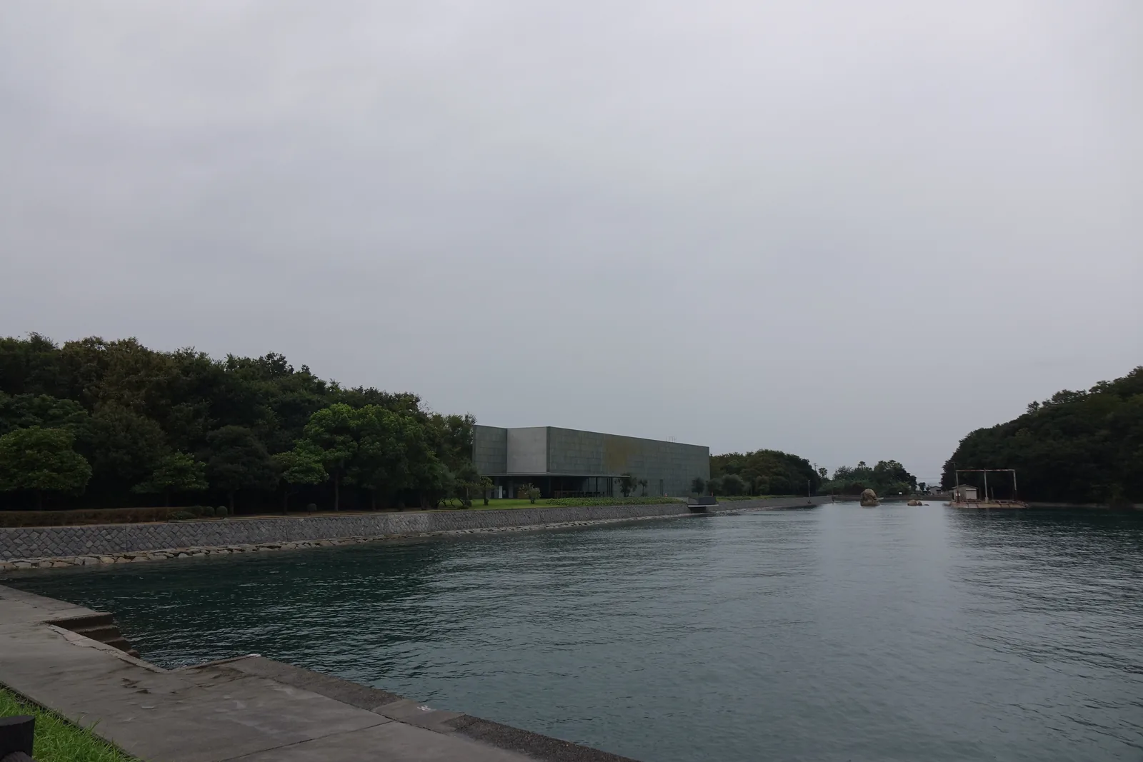 從護岸望向美術館，前景是平靜的海灣與石砌護岸，對岸綠地之後是低矮的灰綠色長方量體，左右兩側是覆滿樹林的小丘