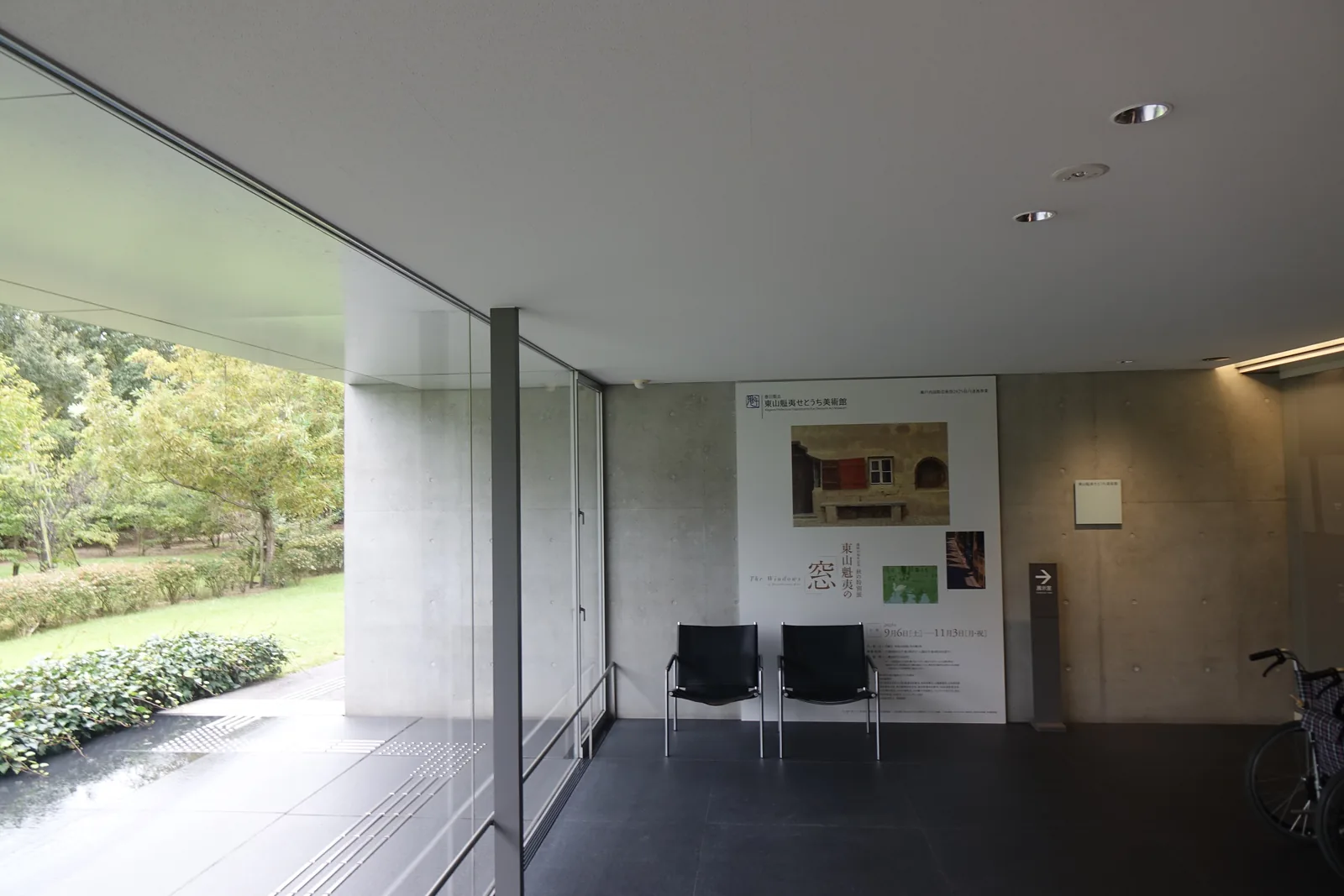 門廳內部，清水混凝土牆上掛著一張展覽海報與一塊小型館名牌，下方是兩張黑色皮椅，左側是整面落地玻璃，窗外是庭園的綠地，地面是深色石材