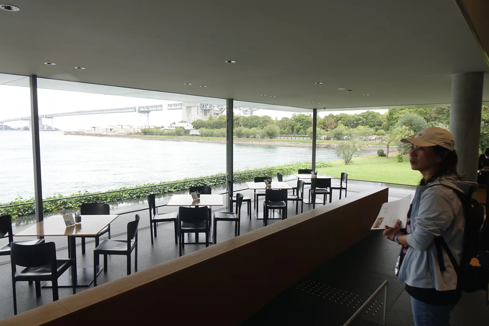 lounge 內部，整面落地玻璃外是灰色的海面與遠處的瀨戶大橋，窗前排著木桌與黑色椅子，右側一位參觀者站在木作矮牆邊望向窗外