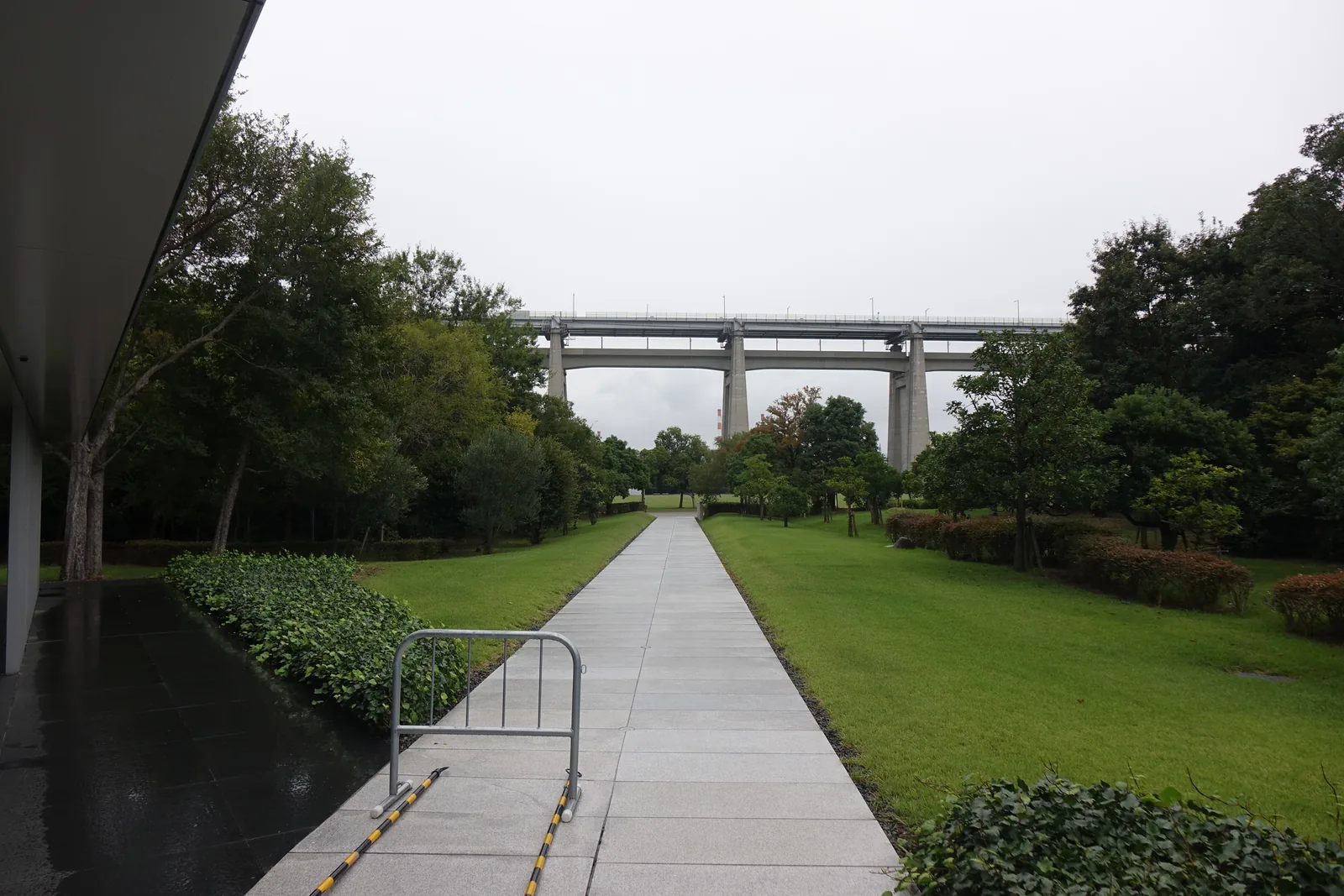 美術館東側的外部空間，左側是建築的屋簷與深色石材的鋪面，一條淺色步道筆直穿過草坪，盡頭是瀨戶大橋的高架引橋與橋墩，兩側是樹木