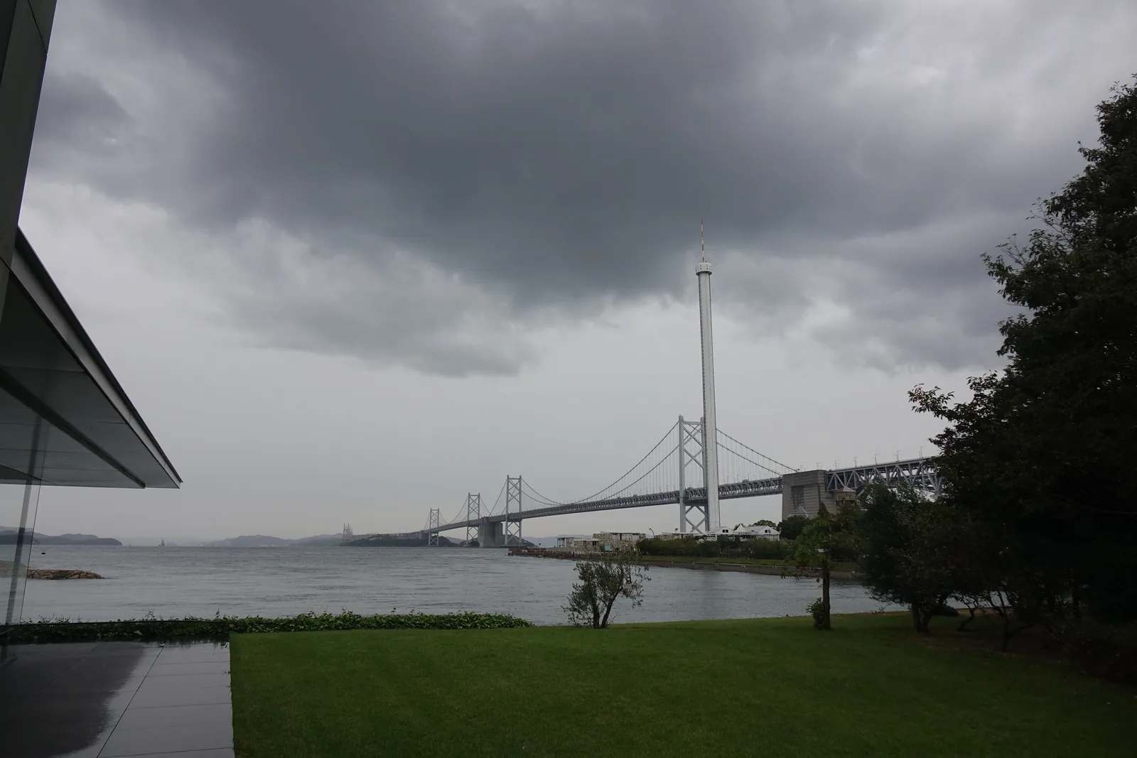 從美術館外部的草坪望向海面，左側是建築的屋簷與玻璃，前景是一片草坪，遠方是瀨戶大橋與白色的瀨戶大橋塔，天空是厚重的雨雲