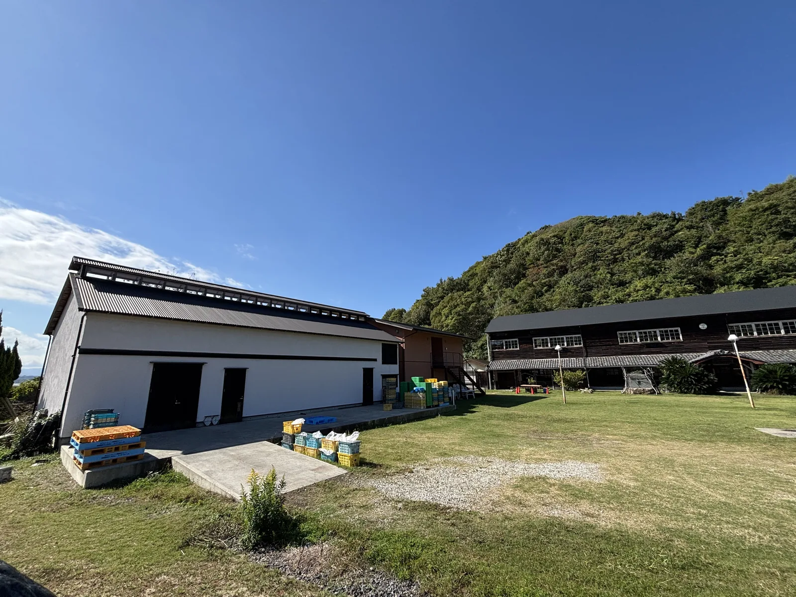 舊宗方小學校的附屬建築與木造校舍，左側是一棟白牆黑瓦的倉庫，右後方是兩層的木造校舍，前景是草地與碎石地面，背後是覆滿樹林的山丘
