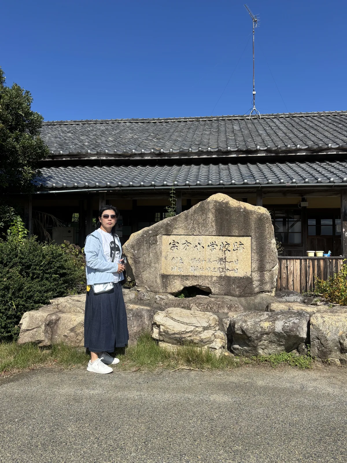 舊宗方小學校遺址的石碑，一塊大石上嵌著寫有學校名稱的石板，背後是兩層的木造校舍與瓦屋頂，左側站著一位同行者