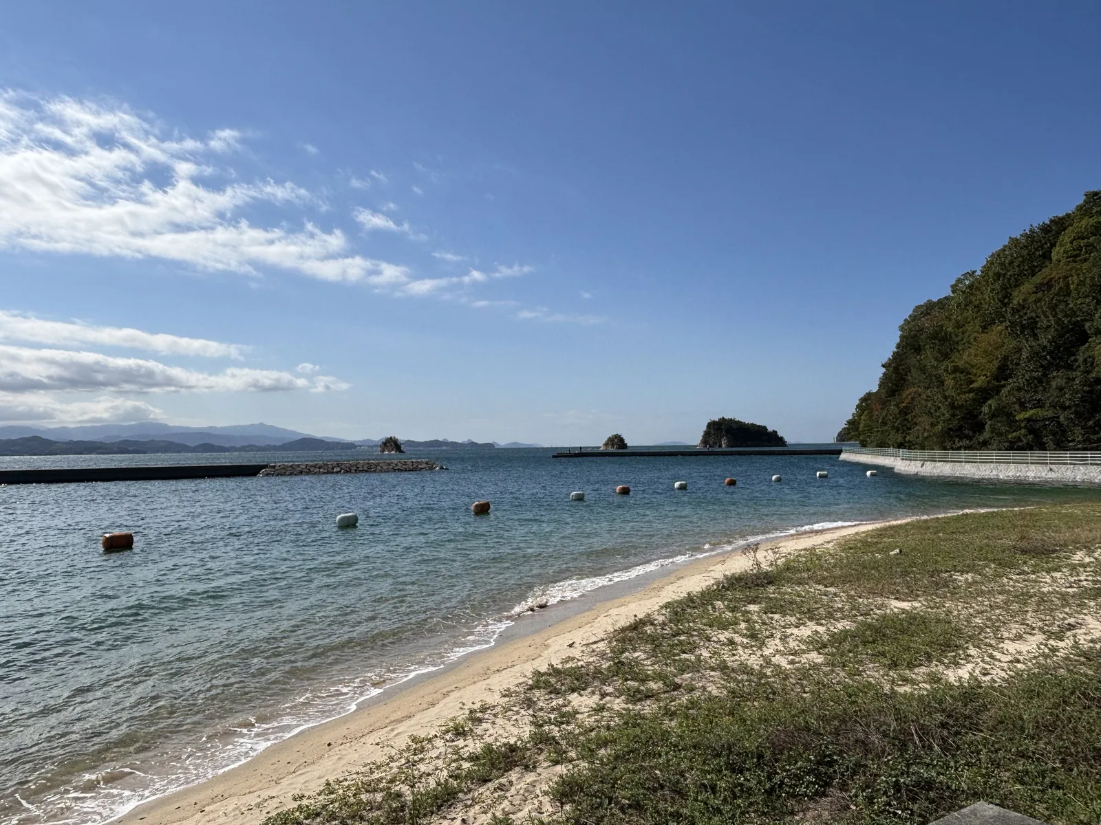 宗方的海灘，前景是長著野草的沙地，中景是弧形的沙灘與平靜的海面，海上漂著浮標，遠處是一道防波堤與幾座小島