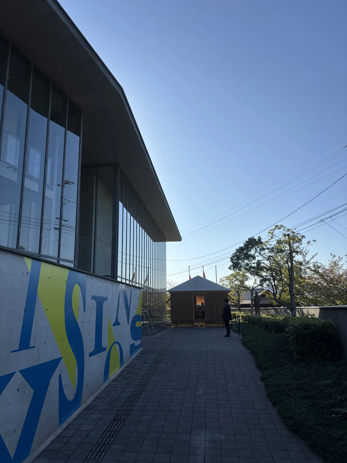 尾道市立美術館新館的西側外觀，左側是整面的鋼框玻璃 screen，上方是向外挑出的薄平屋頂，下方的混凝土牆上漆著藍黃兩色的大型展覽字樣，前方是石材鋪面的通道，通道盡頭是一座紙管構成的小屋