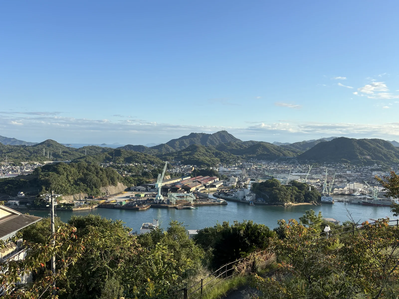 自千光寺公園西側眺望尾道水道，前景是樹林覆蓋的坡地，中景是水道對岸的向島與造船廠的起重機，遠景是一列山稜