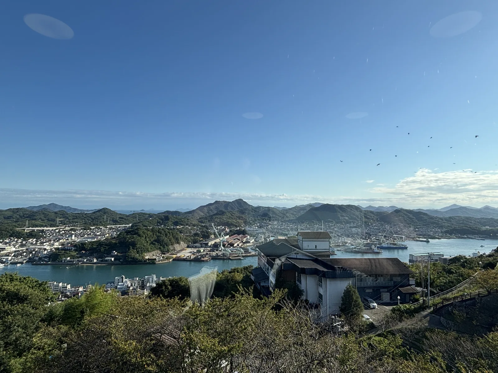 自二樓 lobby 的玻璃窗望向尾道水道，前景是山坡上的樹林與一座白牆的建物，中景是水道與對岸的向島，遠景是一列山稜與晴空