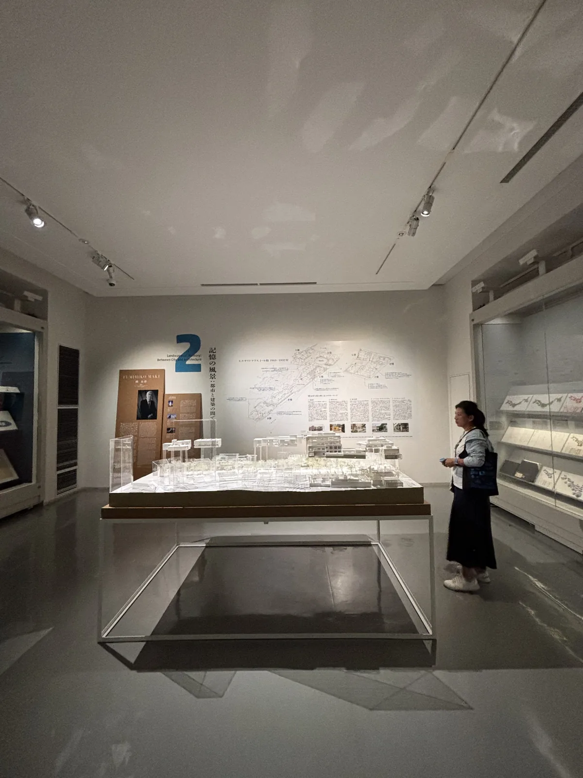 展示室的內部，白色的牆與天花，淺灰色光滑的樓板，中央是一座低矮的展示台，台上放著大型的都市模型，兩側牆面上是展示 case 與圖面，天花上是軌道照明