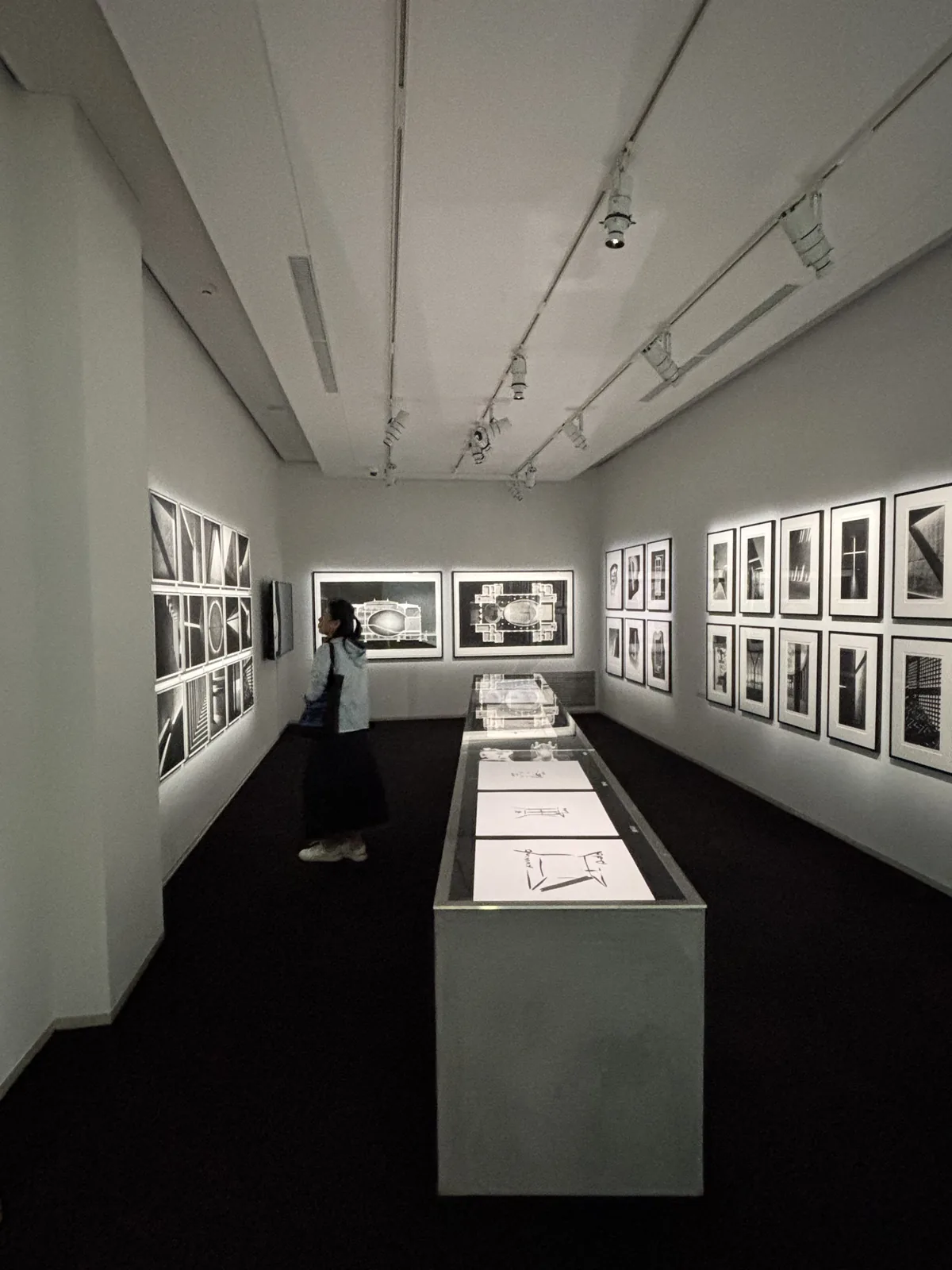 展示室內部，樓板與一側的牆為深色，兩側白牆上整齊排列著黑白建築照片，照片由背後的光照亮，中央是一座長條形的展示台，台面上是建築的速寫