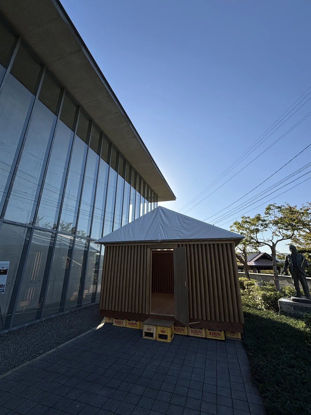 新館西側通道盡頭的紙管小屋，小屋的牆由一根根紙管排列構成，屋頂是白色的膜，基座是一排黃色的塑膠箱，左側是新館的鋼框玻璃 screen，右側是草地與一尊銅像