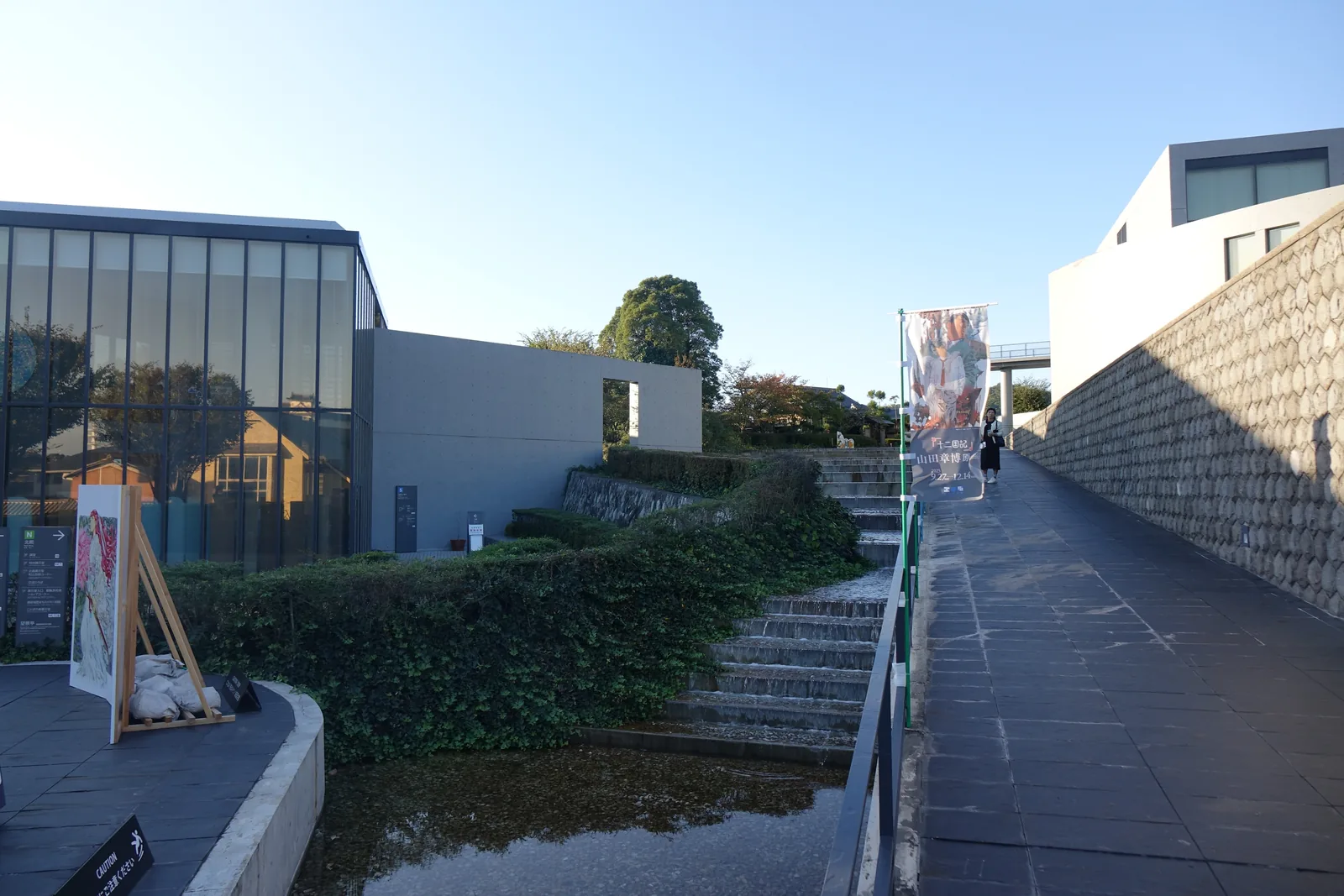 自南館一側通往北館的步道，右側是一道以不規則石塊砌成的石垣，石垣上是修剪過的綠籬，左側是南館的玻璃量體與水盤，步道盡頭是北館的混凝土牆與上行的室外階梯