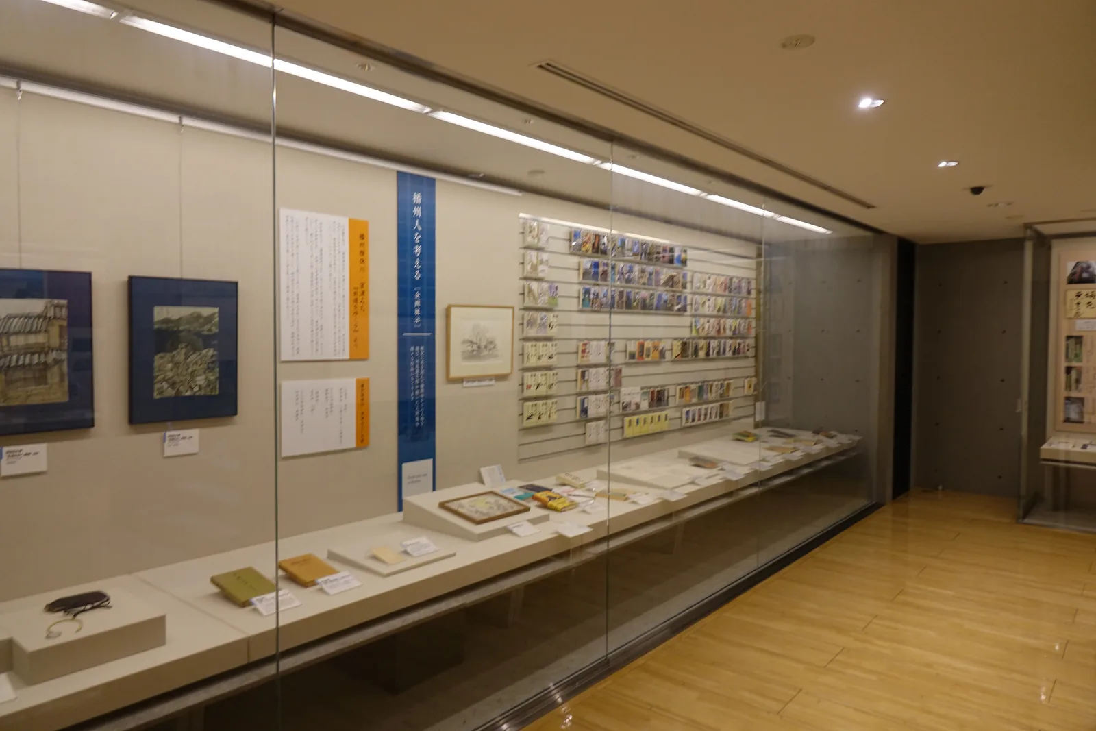 司馬遼太郎紀念室，左側是一整面的玻璃展示櫃，櫃內陳列書籍、原稿與畫作，牆上是直向的說明板，右側是木地板的通道，盡頭的牆面是清水混凝土
