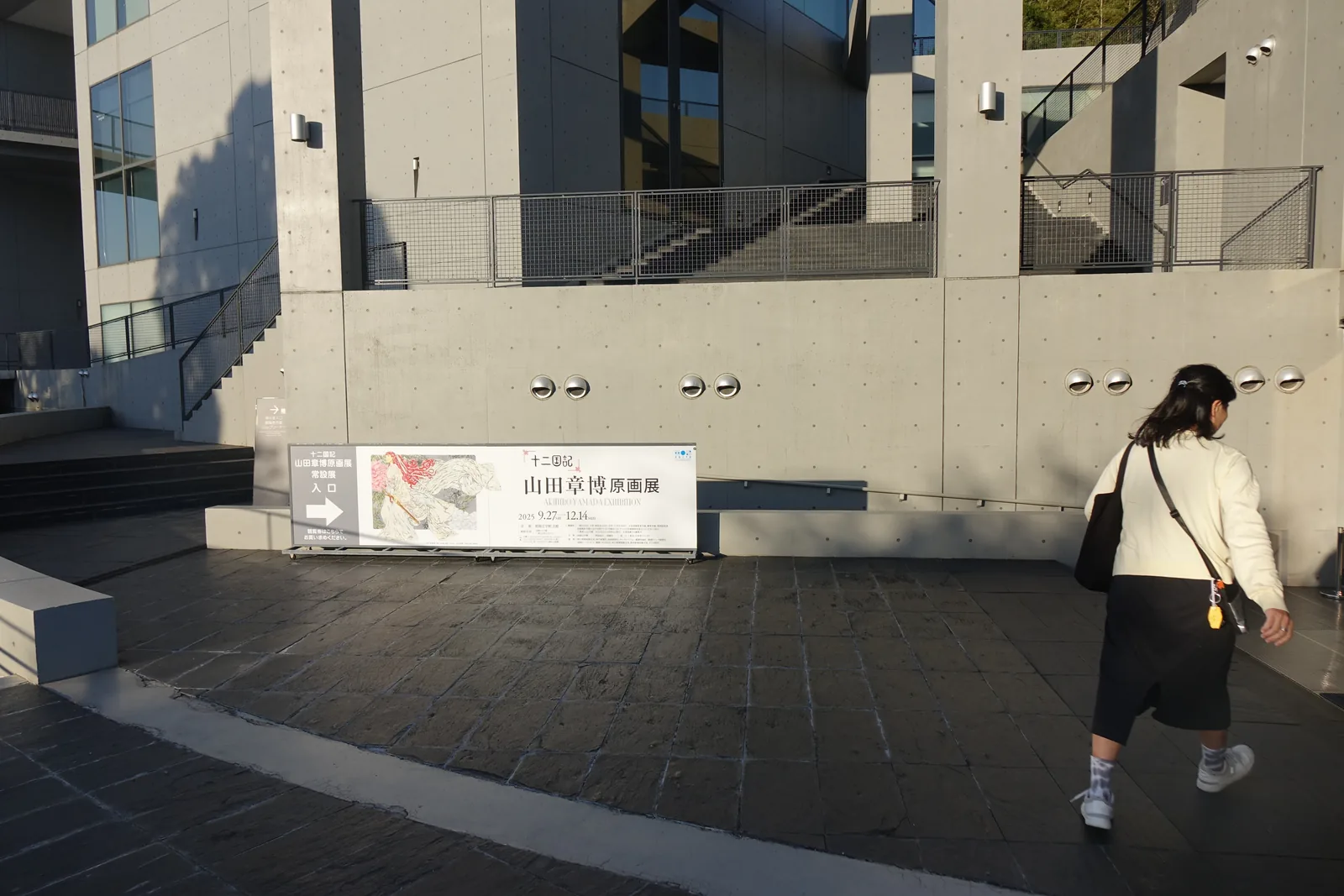 北館前的廣場，地面是深色的石材鋪面，前方是北館的清水混凝土牆面，牆面上有一排圓形的燈具與一面特別展的橫幅看板，左側是向上的室外樓梯與玻璃窗，一位來館者正走過廣場