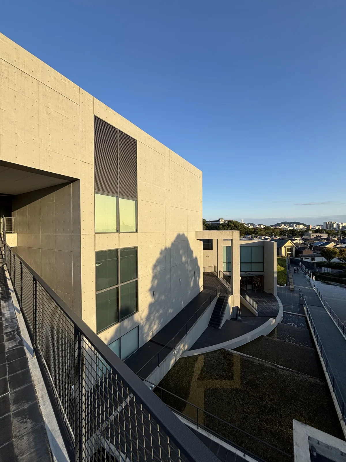 沿著混凝土牆上行的坡道，左側是建築的牆面與玻璃窗，坡道以鋼網欄杆圍起，坡道的盡頭是更高一層的平台與遠方的市街