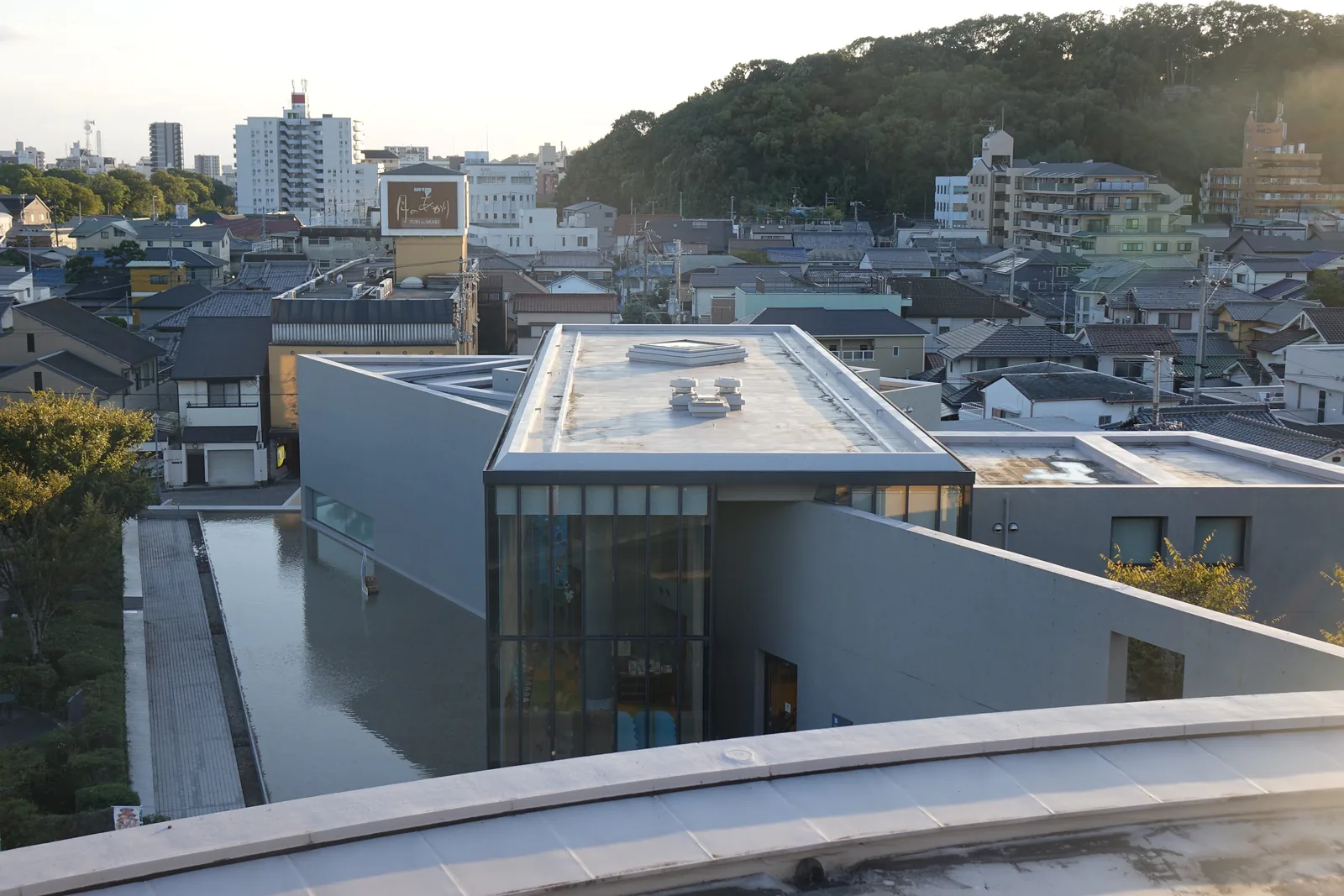 自北館圓筒量體頂部俯瞰南館，前景是圓筒頂部的弧形邊緣，下方是南館的長方形量體，量體的一端是玻璃立面，屋頂平而淺色，左側是一道細長的水盤，遠處是市街與一座覆滿樹林的山