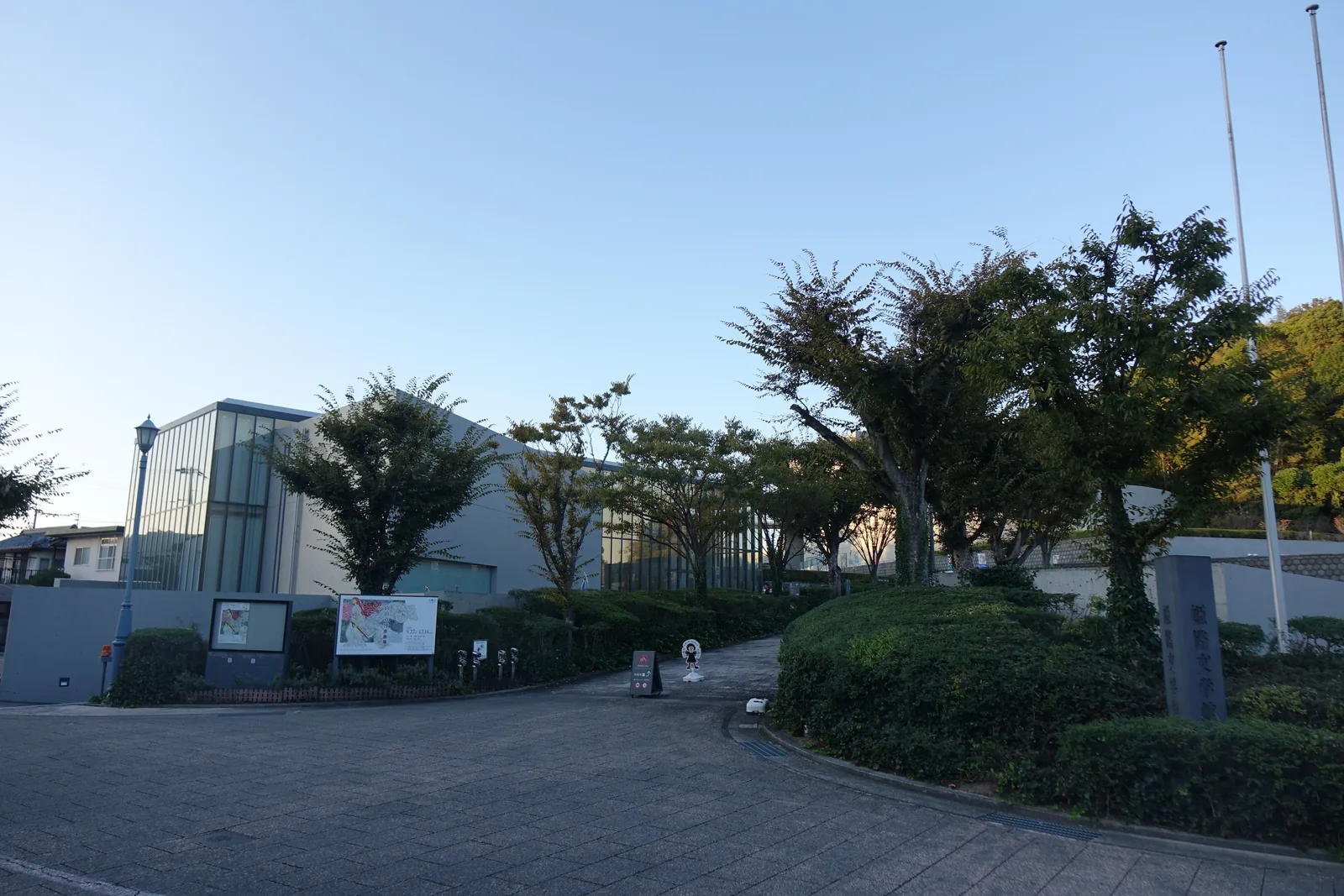 文學館臨街的正門一帶，前景是鋪石的車道與修剪成圓形的綠籬，中景是成排的樹與兩根旗桿，樹後是南館的玻璃量體與館名石碑