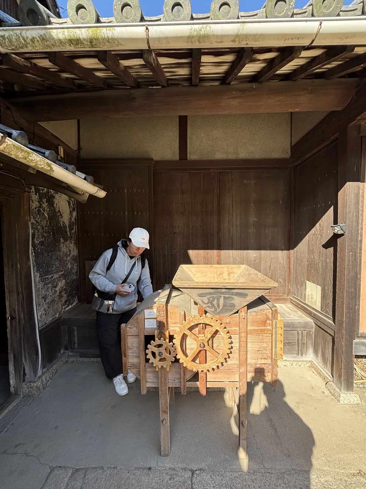 民家入口的簷下，屋簷的椽木與瓦片之下擺著一具木製的農具，側面裝著兩個大小不一的木齒輪，背後是木板牆與門，一位參觀者站在一旁
