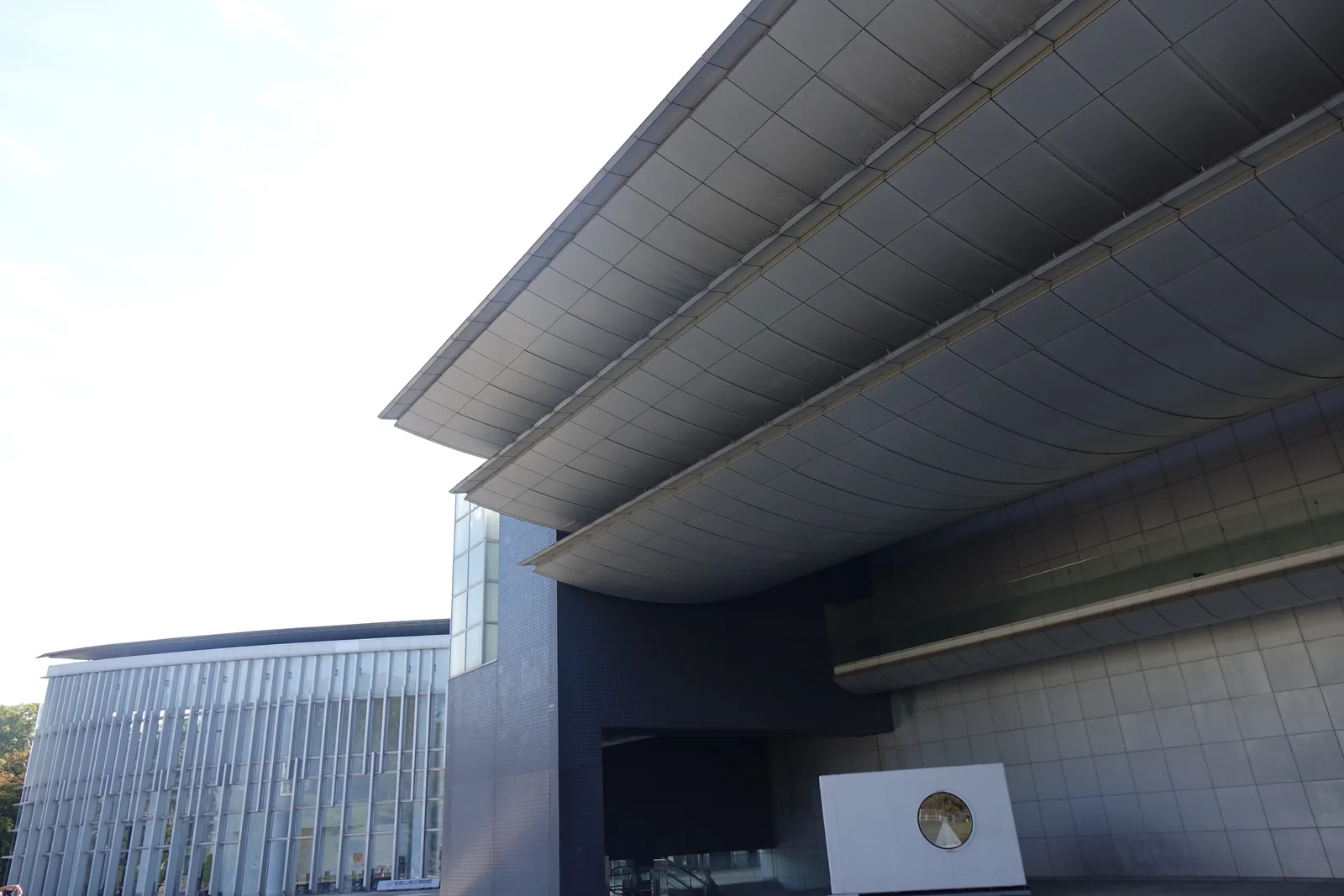 美術館的金屬庇自上方伸出，畫面右側是美術館深色的量體與多層的庇，左側遠處是博物館以白色直向百葉構成的弧形立面，中央是一面嵌著圓形浮雕的白色石板