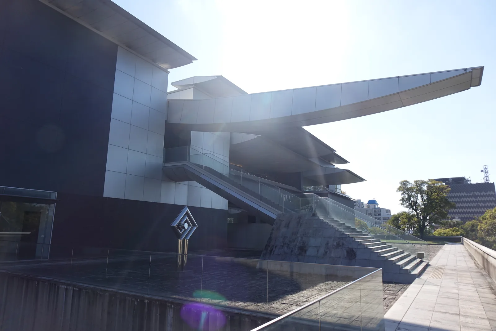 美術館側面的長庇，一道細長的金屬庇自建築側面伸出，前緣微微上翹，下方是沿著立面上行的室外樓梯與玻璃扶手，左側是深色的量體與一件菱形的金屬雕塑，右側是石材鋪面的步道