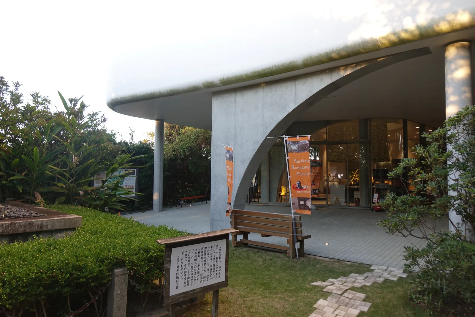 南方熊楠紀念館新館的一樓外部，一道清水混凝土的半拱自地面升起，上方是邊緣圓滑的白色樓板，樓板頂部長著草，右側是一根細的圓柱，拱與柱之後是玻璃圍出的大廳，前景是草地、長椅與一塊說明板