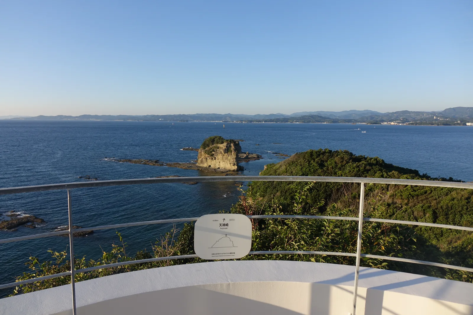 自新館屋頂所見的海岬，前景是覆滿常綠樹林的山脊，山腳是岩礁，海中立著一座頂上長樹的岩島，遠方是對岸的山與海岸線