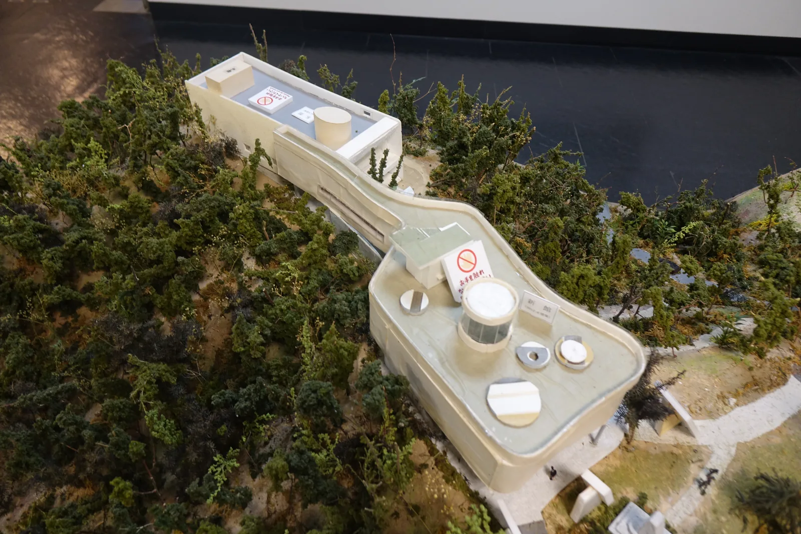 番所山的地形模型，白色的建築量體沿著山頂細長地排開，四周以深綠色的模型樹表示森林，山腰有數條灰色的步道與車道