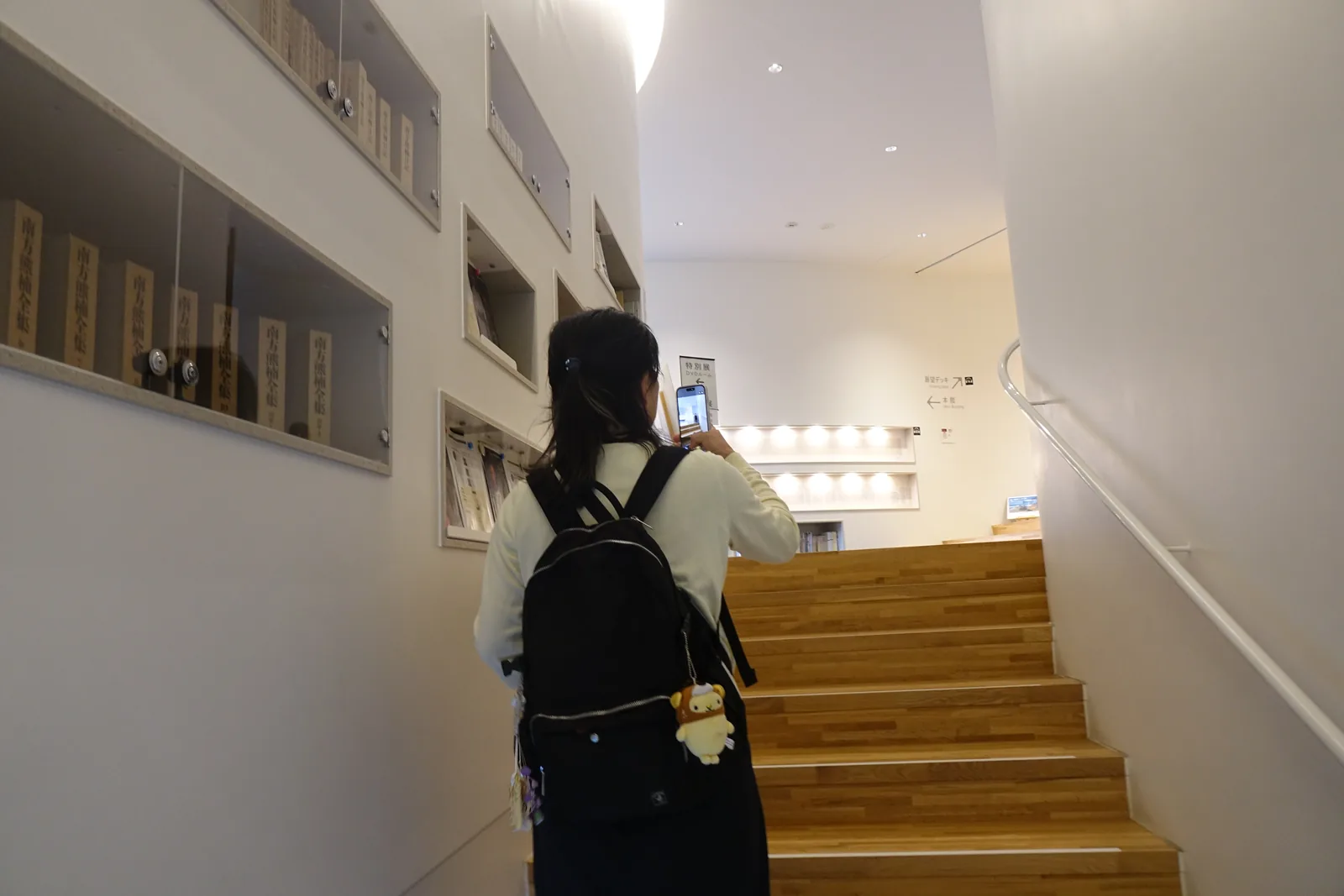 通往二樓的樓梯，木質的踏面向上延伸，左側的白牆上開著一列方形的壁龕，壁龕內陳列書籍與資料，樓梯盡頭是更高的白牆與燈光，一位來館者正在樓梯上