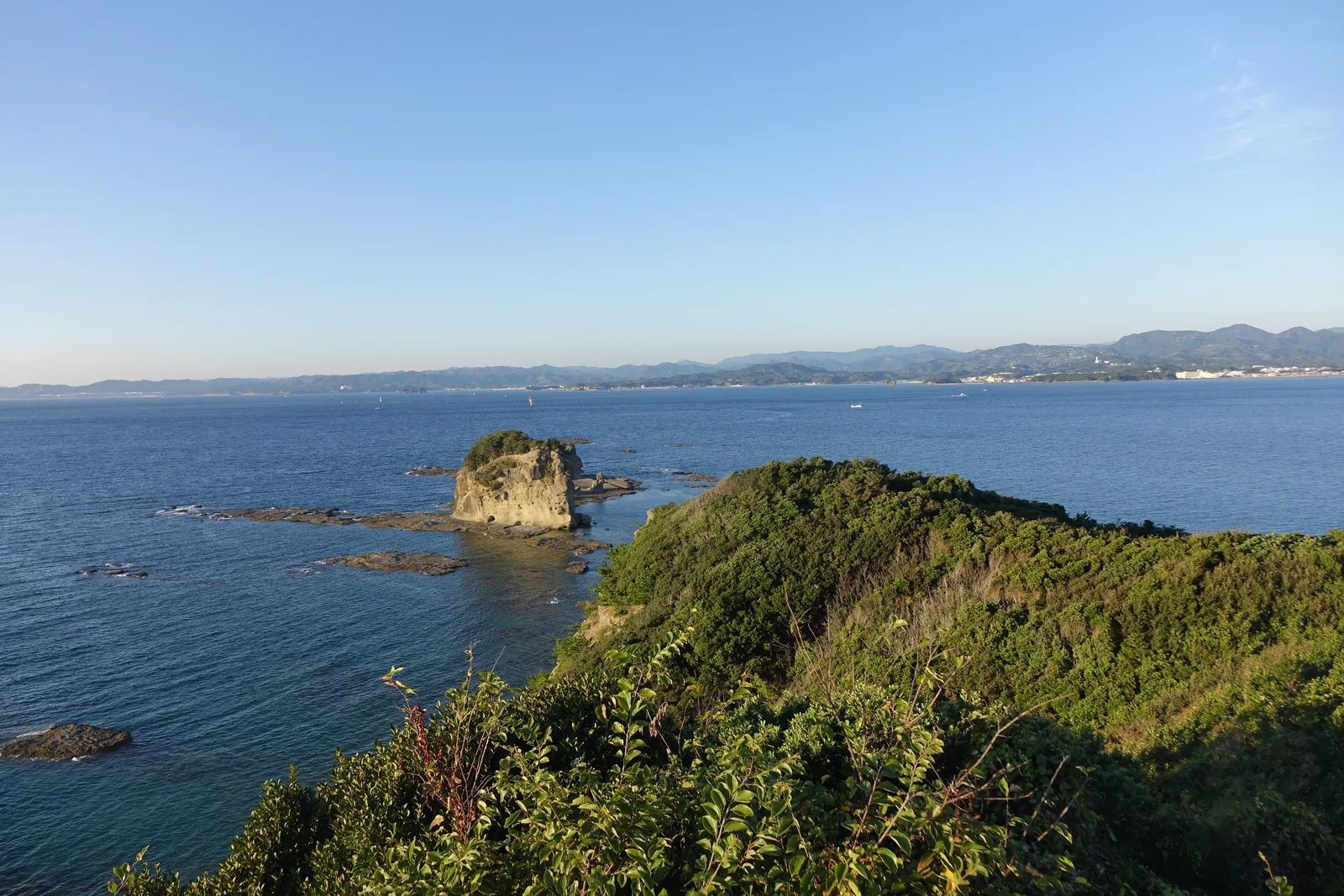 自新館屋頂望向本館，前景是覆滿樹林的山坡，中景是本館白色的長方形量體與屋頂上的圓筒形塔屋，背景是海與對岸的山
