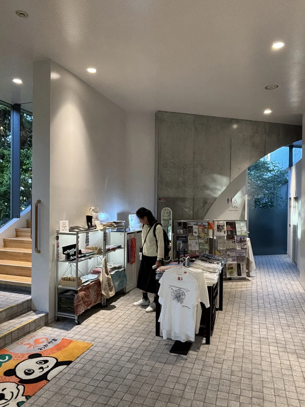 新館一樓入口大廳一角的商店，左側是陳列書籍與雜貨的商品架，中央是吊掛的 T 恤，地面是淺色的方形磁磚，後方是通往二樓的樓梯與清水混凝土的牆，一位來館者正在選購