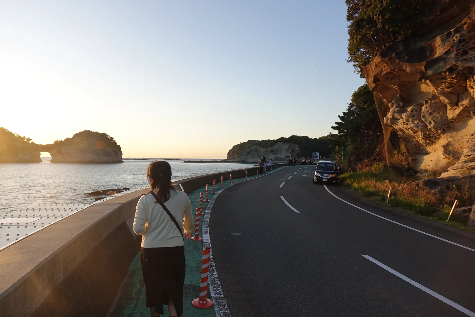 臨海浦的沿岸道路，右側是岩壁與車道，左側是海堤，海中是中央穿洞的圓月島，一位行人沿著海堤行走，天色是傍晚