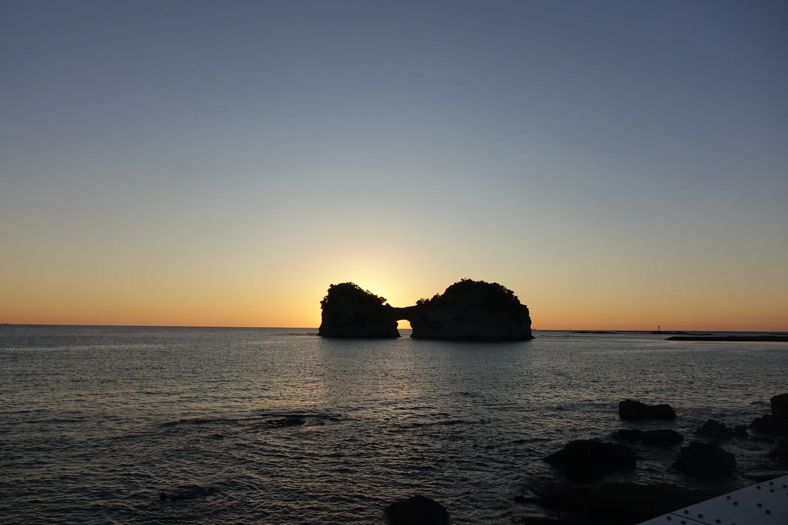 圓月島的日落，太陽落在中央穿洞的岩島左側的縫隙之間，海面映著一道光，前景是海岸的岩石