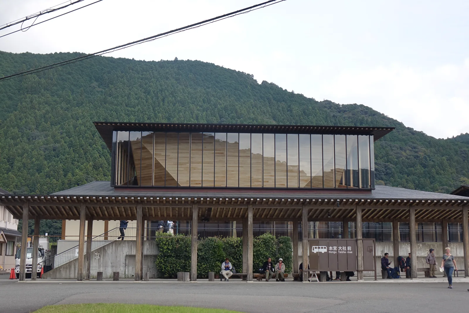 世界遺產熊野本宮館臨國道的立面，前方是一道以方形木柱撐起的低矮簷廊與巴士站牌，簷廊上方是一座整面玻璃包覆、內側有縱向木材的較高量體，背景是覆滿杉林的山