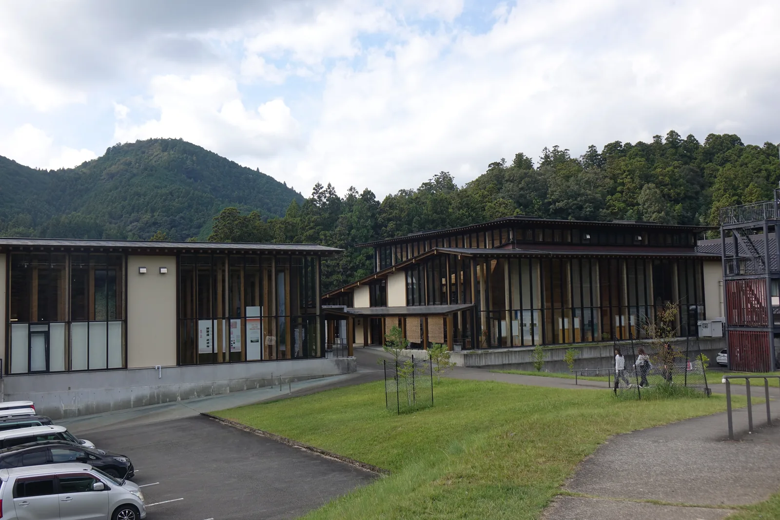 自熊野川堤防回望熊野本宮館，兩棟以玻璃與木框構成立面的平屋並排在一片草坡之上，建築底部是一道混凝土基座，前景是停車場與草坡間的步道，背景是杉林覆蓋的山
