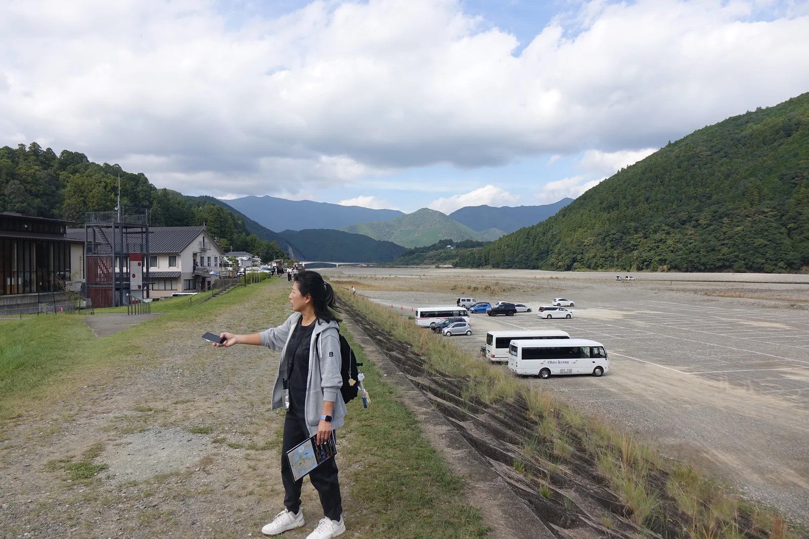 熊野川堤防上的視野，左側是熊野本宮館與一座塔狀的鋼構，中央是寬闊的河灘砂礫地，上面停著巴士與汽車，背景是層疊的山稜，前景是一位站在堤防步道上的人