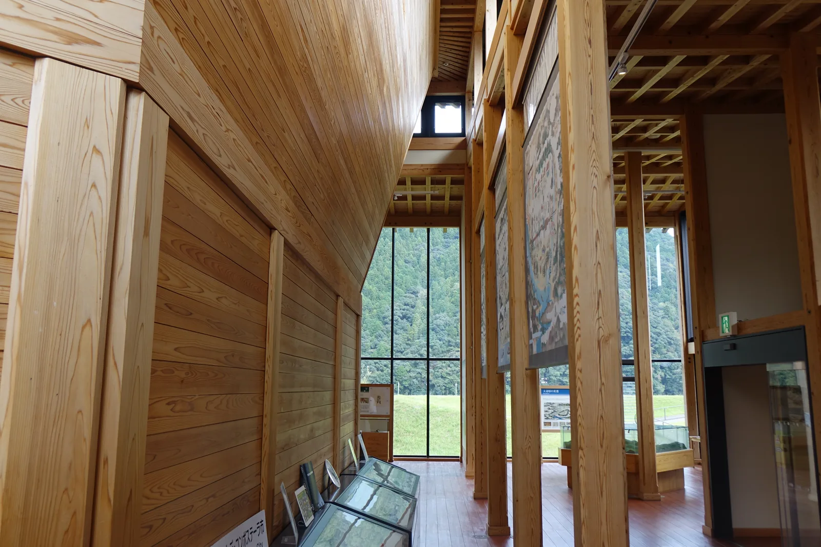 展示空間沿外牆的通道，左側是以杉板構成、向上傾斜的牆面，右側是一排方形木柱與懸掛的掛軸畫，天花是木材組成的細長格子，盡端是整面玻璃，玻璃外是草地與杉林