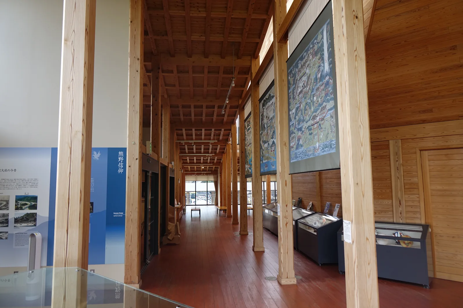 長向的展示空間，左右兩排方形杉木柱向遠處延伸，形成一道柱廊，天花是細長的木格子，右側柱間懸掛著大幅的繪畫複製，地面是紅褐色的木地板，沿牆排著展示櫃，盡端是明亮的玻璃面