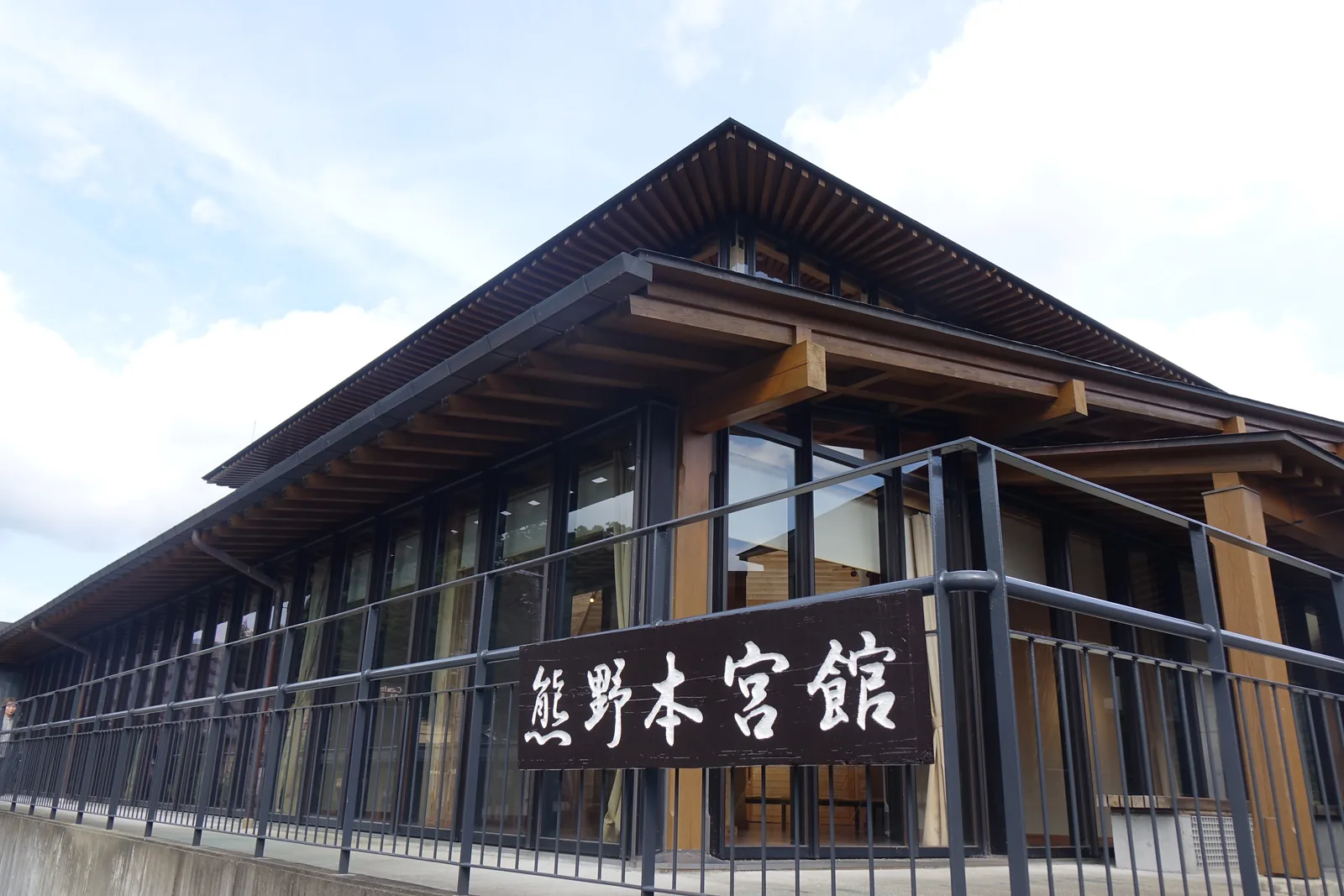 熊野本宮館的轉角，深出的屋簷下露出成排的木樑端部與斜撐，屋簷下是整面以木框分割的玻璃，前方是一道鋼製欄杆與混凝土基座，欄杆上掛著寫有館名的木製招牌