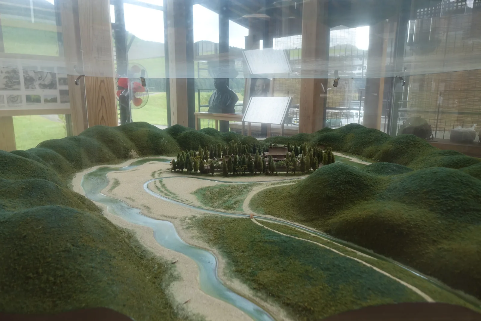 熊野本宮大社舊社地大齋原的立體模型，綠色的山丘之間，三條河道環繞一塊中洲，中洲上是一片樹林，背景是展示空間的玻璃面與木柱