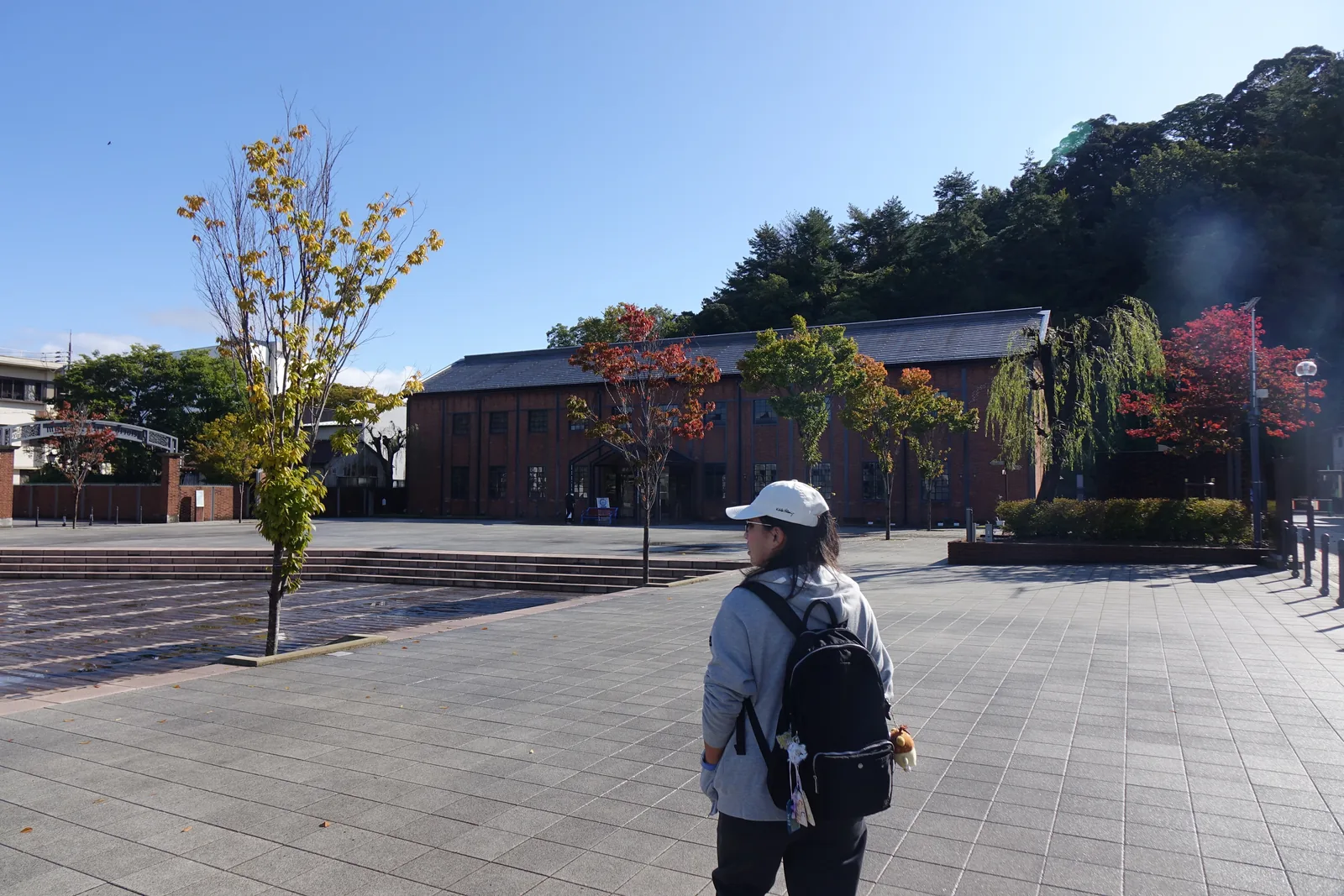 博物館前的廣場，大面積的石材鋪面上站著一位戴帽背包的人，左側是一株秋季變色的小樹，遠處是一棟瓦葺屋頂的低矮紅磚建築與一排紅葉的樹，背景是山