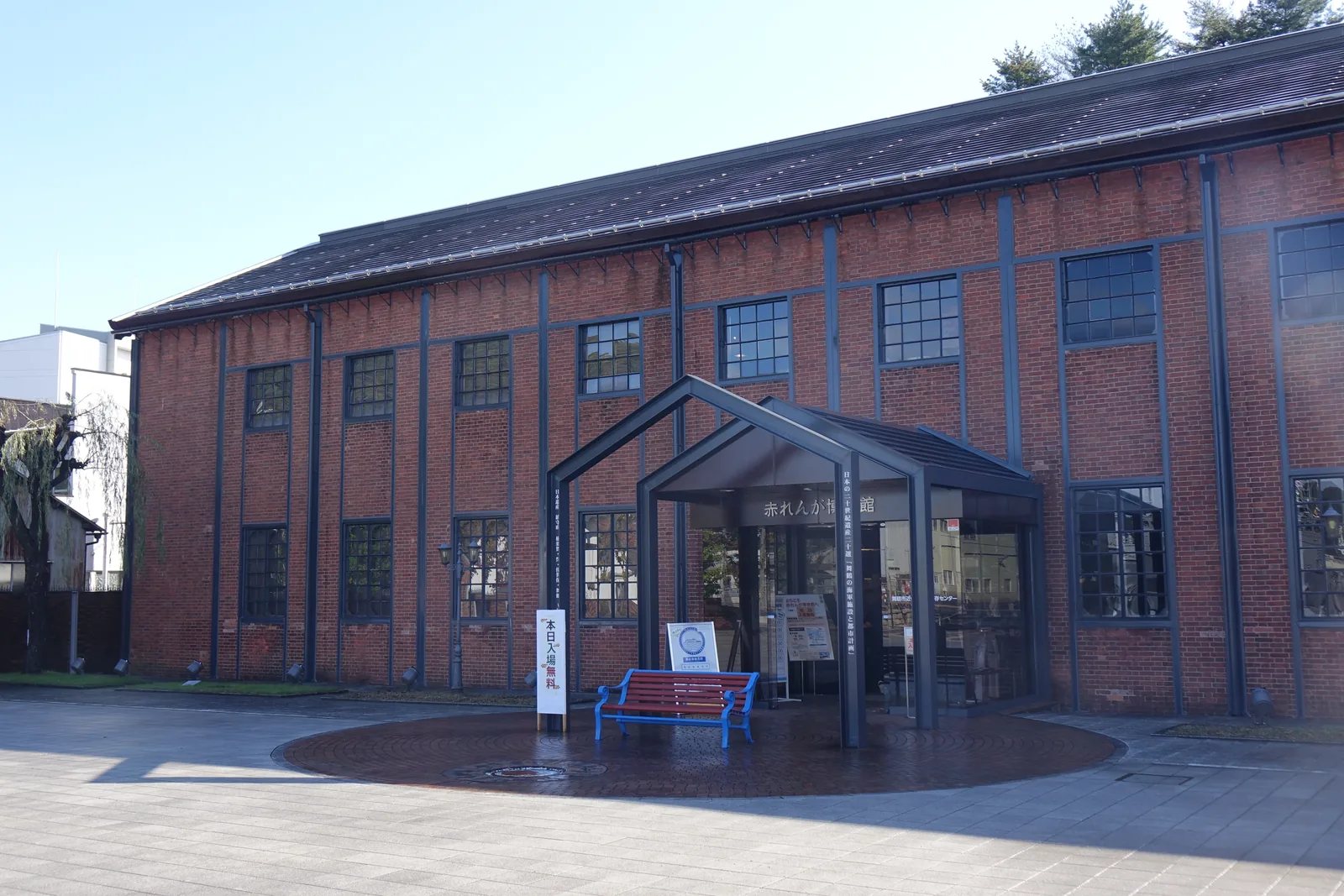 紅磚博物館的正面，一棟兩層的紅磚長向建築，立面以壁柱分割，每跨上下各一扇方窗，入口前方是一座深色鋼構件組成的雙山形門廊，門廊前擺著一張紅色長椅，前方是圓形的磚鋪地面