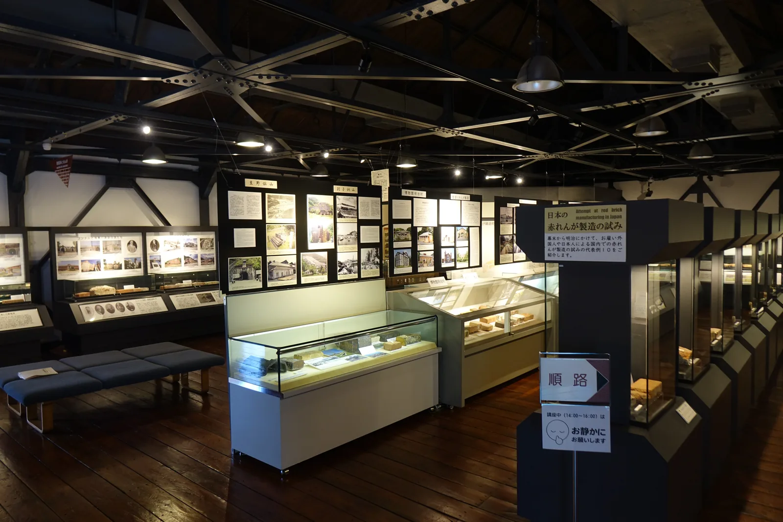 紅磚博物館二樓的展示空間，深色的木地板上排著展示櫃與展示牆，頂部是深色的鋼桁架與木製屋面板，吊燈從屋架垂下，牆上掛著說明板，右側立著一塊順路指示牌