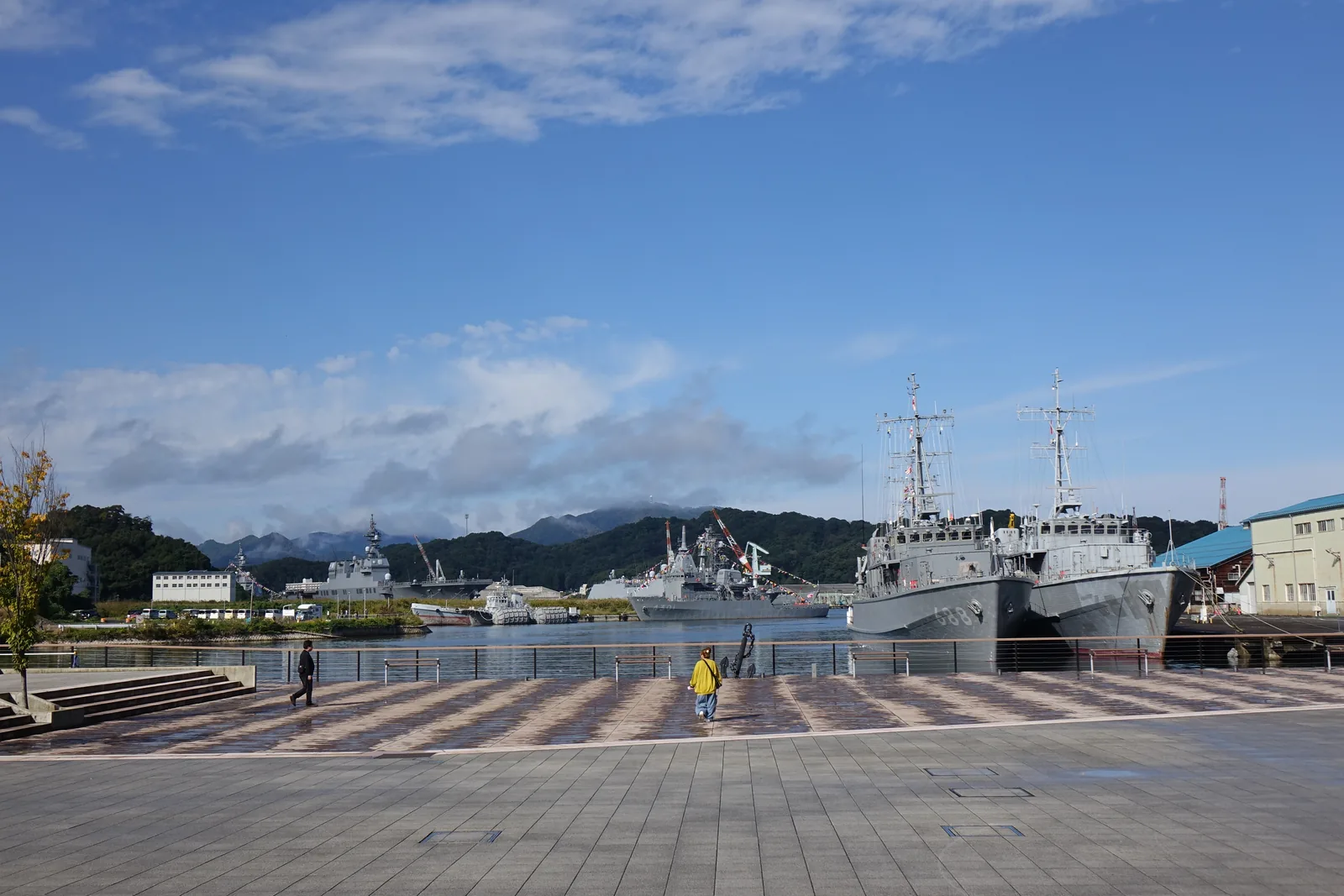 紅磚博物館前廣場的港邊，大面積的石材鋪面延伸到海邊的欄杆，對岸停泊著兩艘灰色的艦艇，背景是山與白雲，廣場上有幾位散步的人