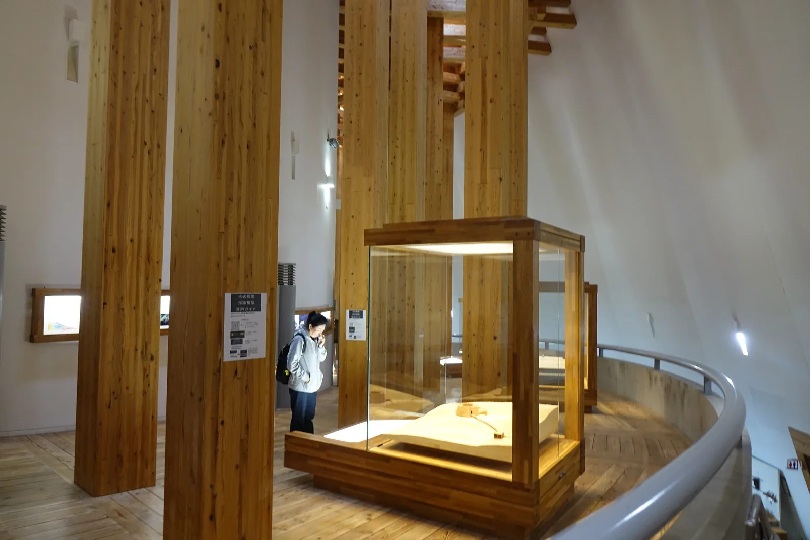 環狀展示空間的一角，前景是兩根粗大的集成材柱與一座木框玻璃展示櫃，右側是沿曲面延伸的扶手與木地板，一位參觀者站在展示櫃旁，背景是白色的牆與小窗