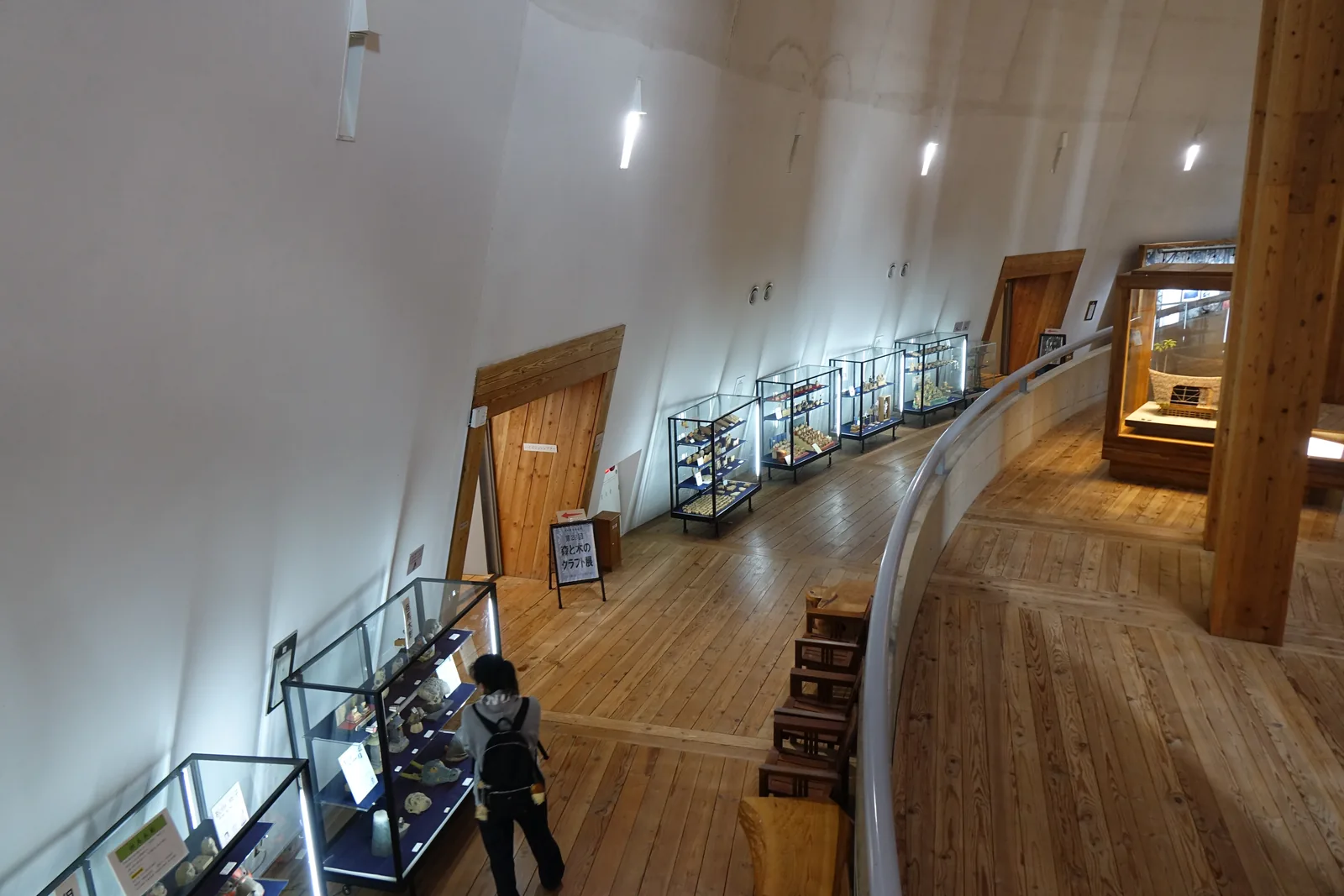 自上方俯視環狀展示空間，木地板沿著曲面延伸，左側是一排展示櫃與一扇木門，右側是曲面的扶手與木柱，一位參觀者在展示櫃前，牆面是白色的曲面與壁燈