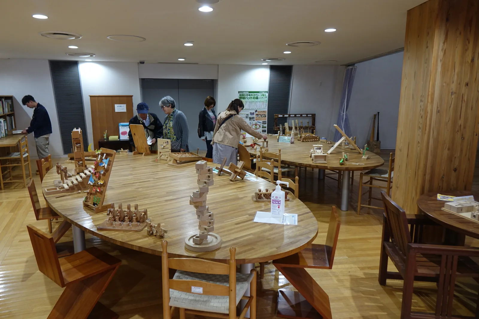 一間房間裡的大型木桌，桌上擺著組木的作品與木製玩具，幾位參觀者圍在桌邊，背景是白色的牆、一根粗大的木柱與幾張木椅