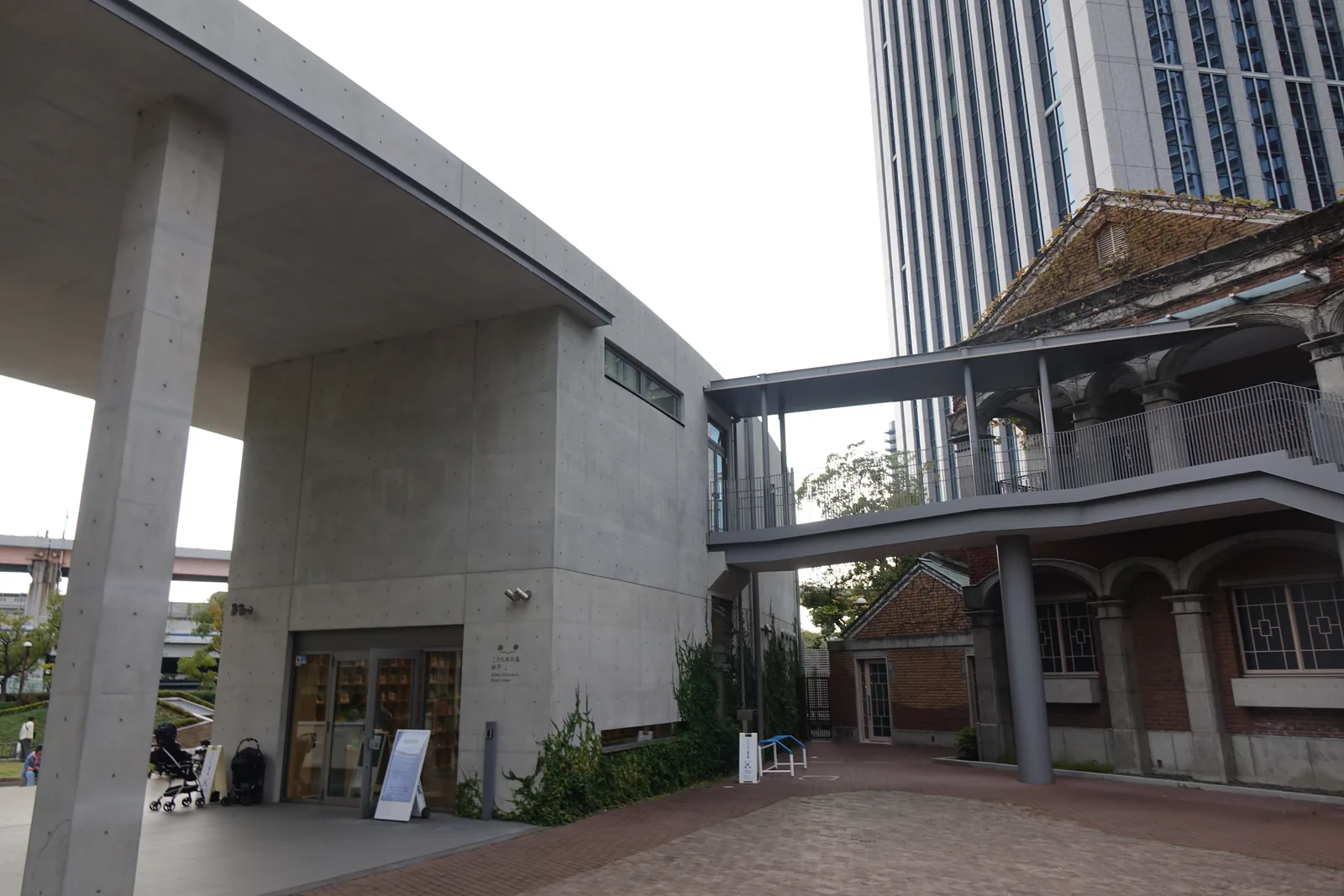 兒童之書森林・神戶的入口，左側是一片清水混凝土的平屋頂以細柱承在端部，屋頂下是深色框的玻璃門與書架，右側一道鋼構連絡橋自二樓伸出，接上一棟紅磚拱廊的二層建築，背景是高層辦公大樓