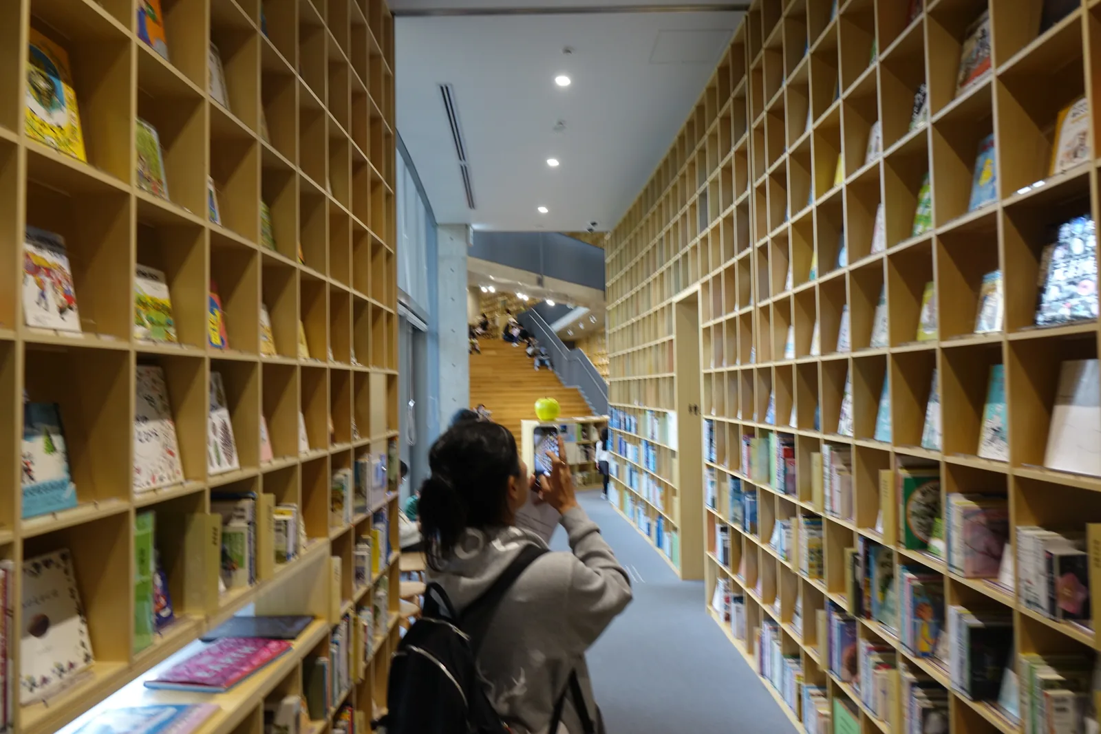 一樓的書架走道，兩側都是自地面到天花的原木色格狀書架，上段的書以封面朝外陳列，走道盡頭可見大階梯與一顆綠色的蘋果