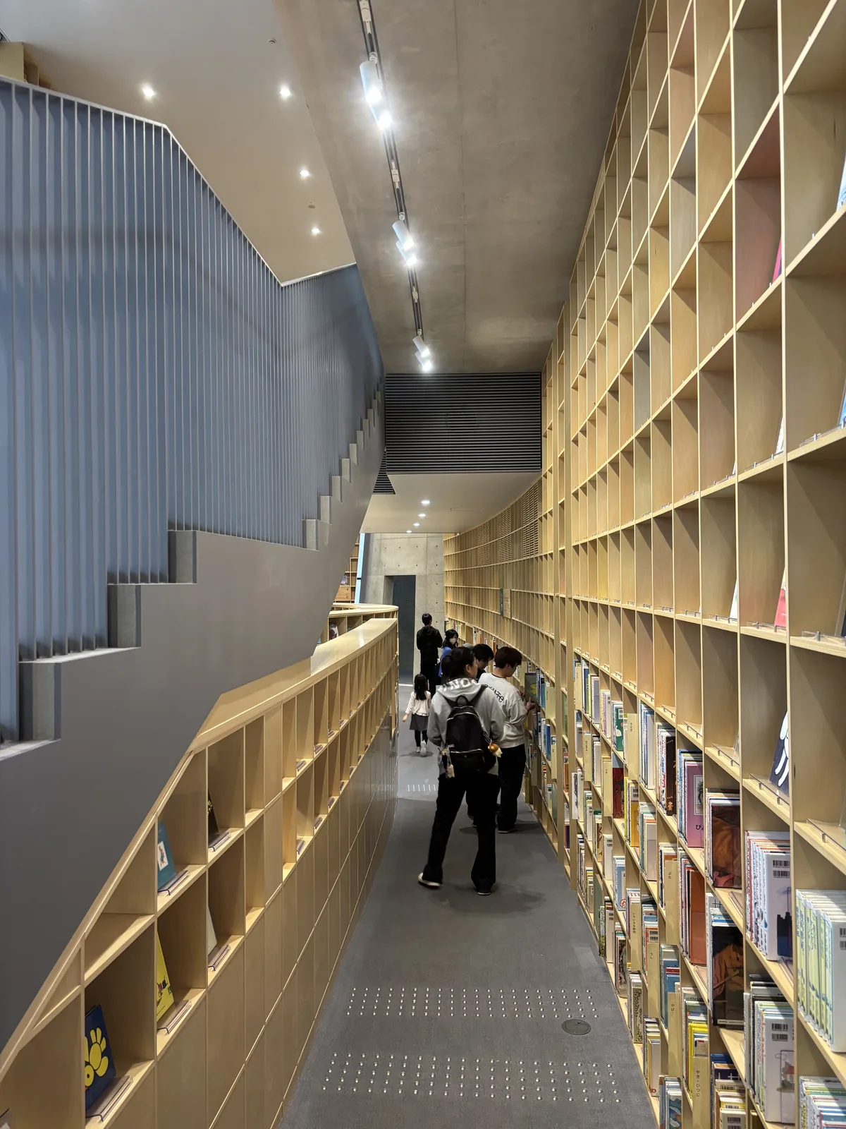 沿著大階梯側邊延伸的通路，左側是階梯的鋼板欄杆與階梯下的矮書架，右側是自地面到天花的書架，地面鋪著灰色地毯與導盲磚，遠處有數位來館者