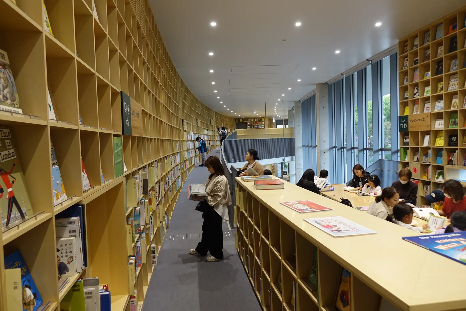 二樓的閱覽區，左側是沿著彎曲的牆面延伸的書架，右側是一道長形的木製閱覽桌，桌下是書架，桌旁坐著許多大人與兒童，窗外是樹木