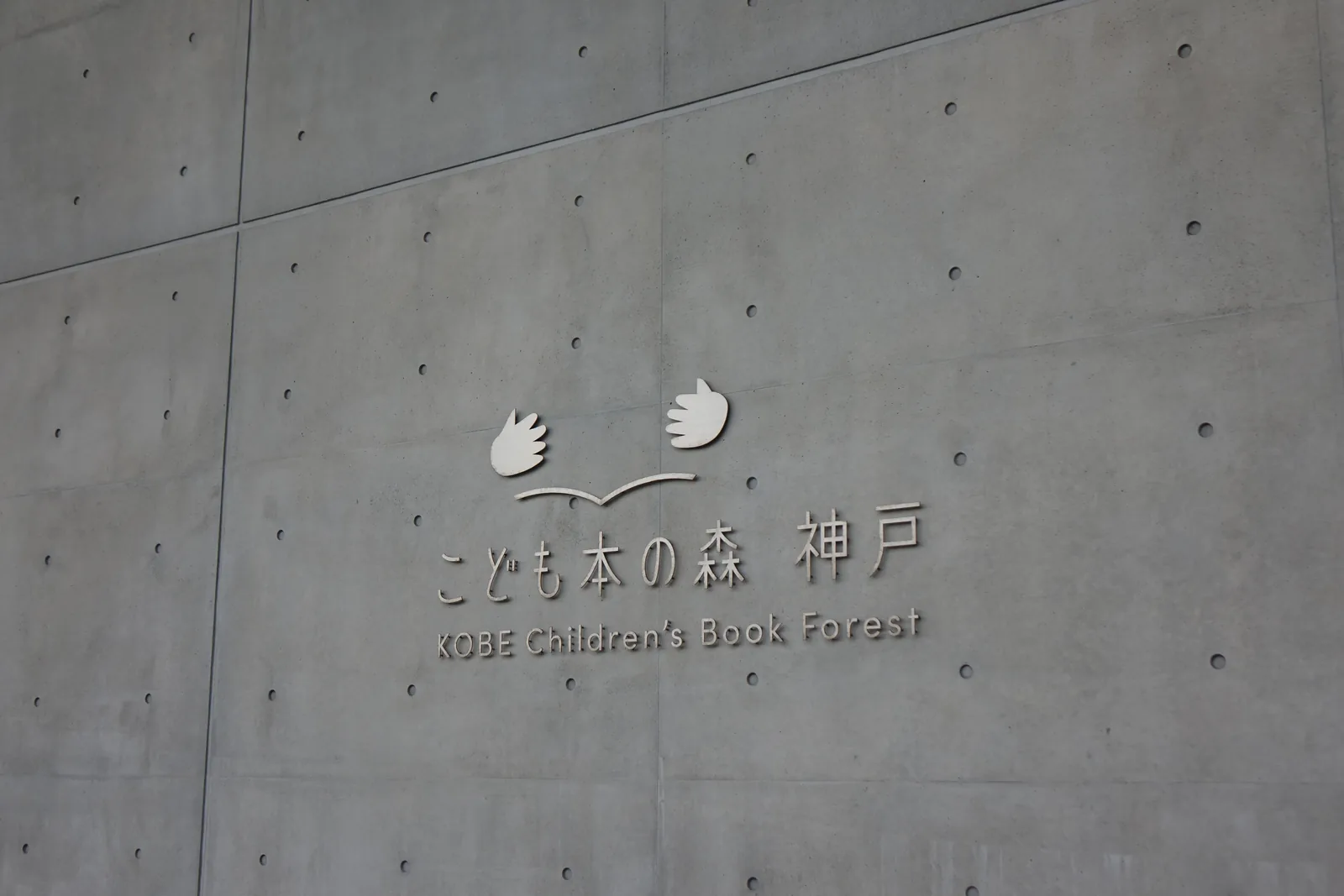 清水混凝土外牆上的館名，金屬字寫著館名與英文名，其上是兩隻手捧著一本書的標誌，牆面留著模板分割線與螺栓孔